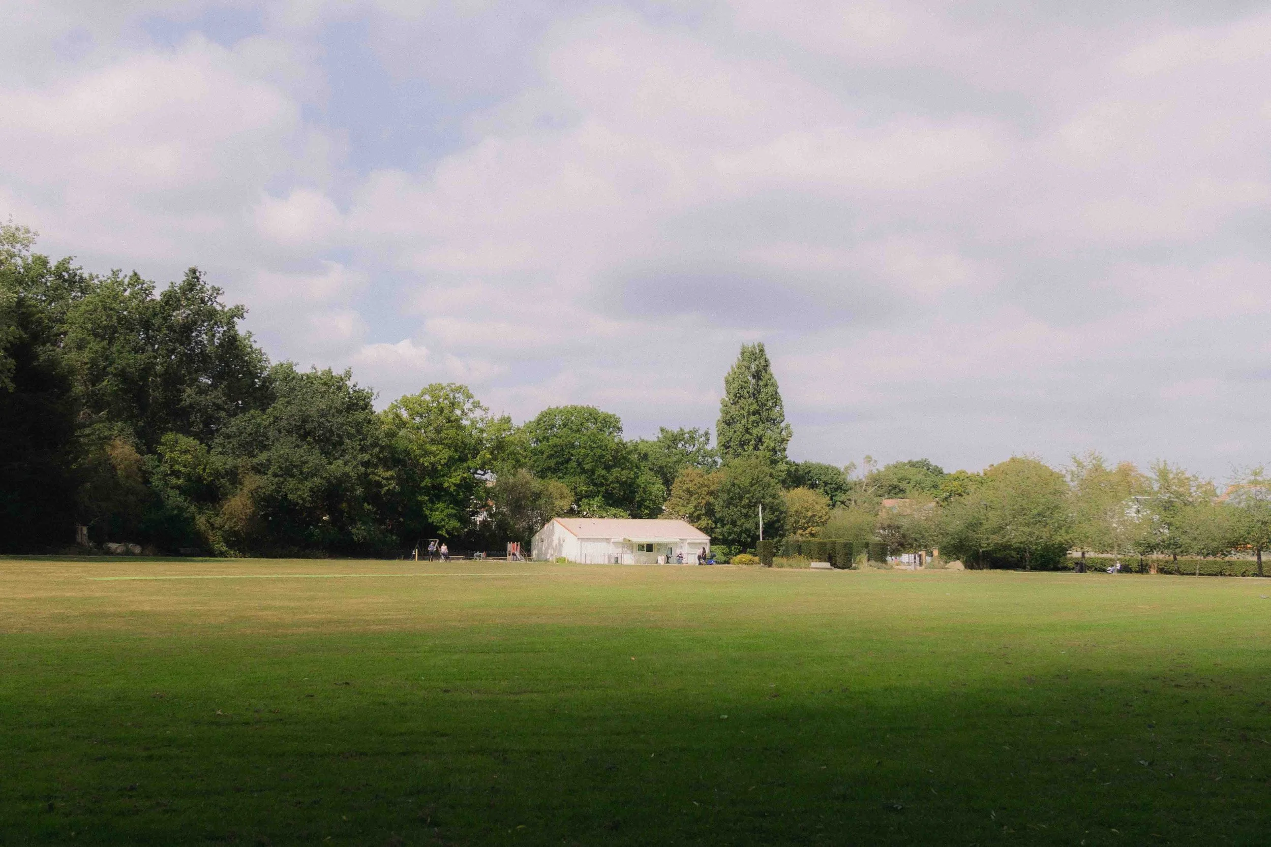 Open grassy field with a small white building and trees in the background under a partly cloudy sky.