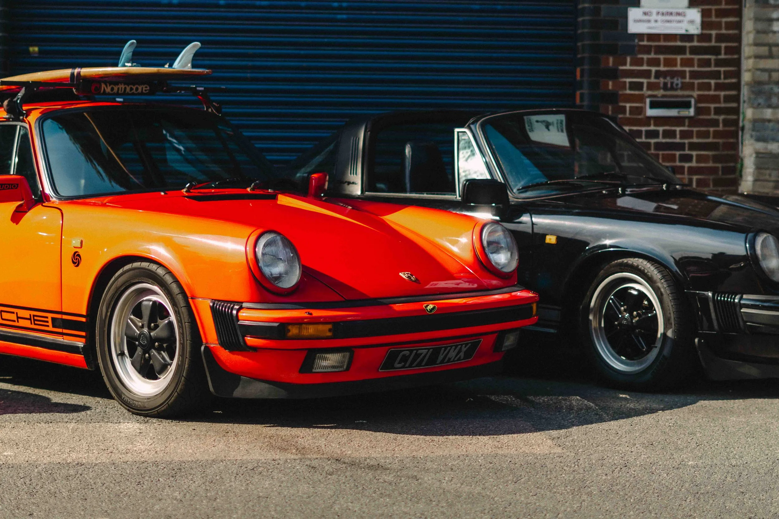 Two classic Porsche cars parked side by side on the street. One is bright orange with black accents, and the other is black with a silver roof, both with round headlights and retro design.
