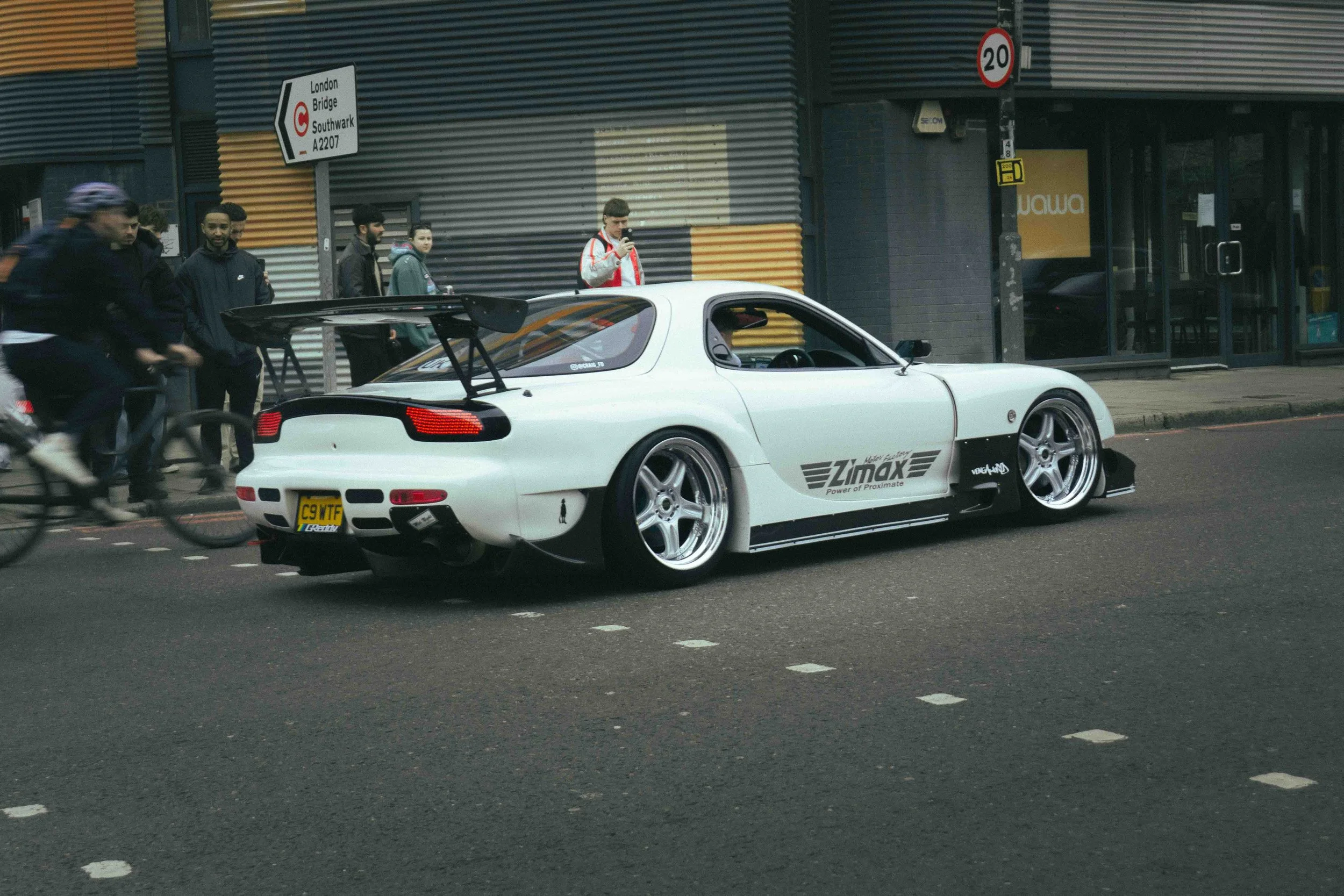A white sports car with black and gray decals, large rear wing, lowered stance, and aftermarket wheels parked on the street near a group of pedestrians and cyclists.