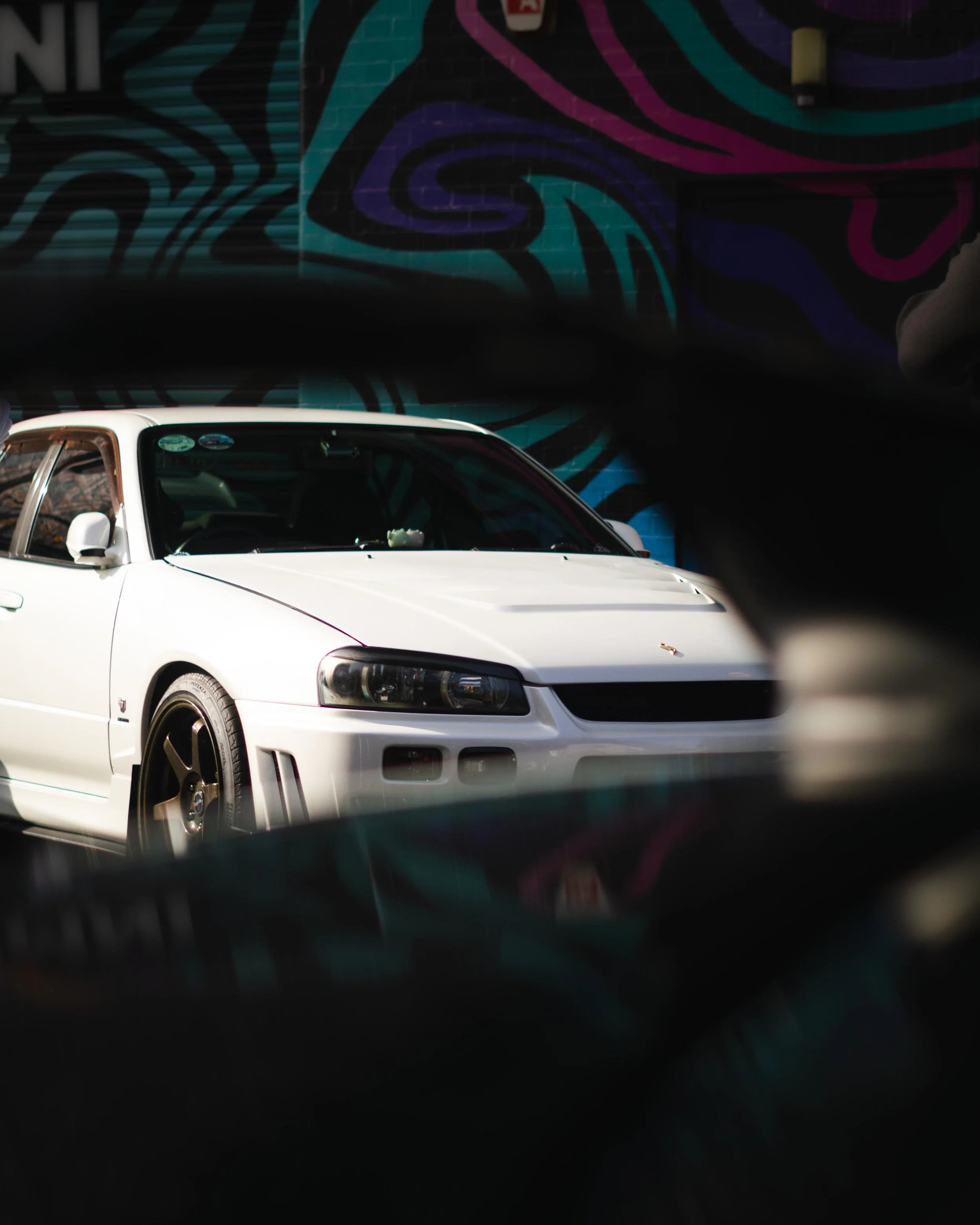 A white sports car parked in front of a colorful, abstract, mural wall with black, teal, purple, and pink lines.