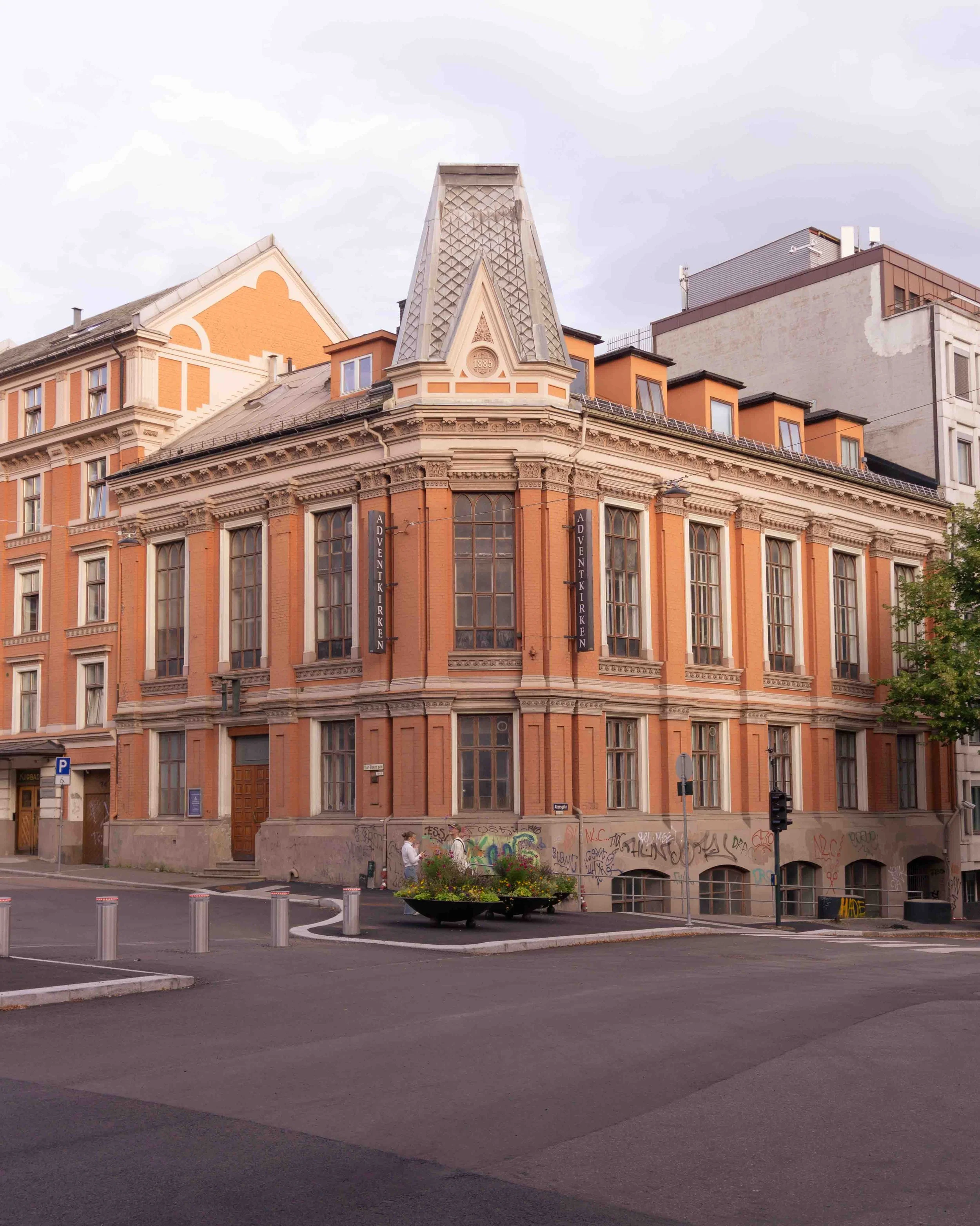 A historic orange brick building with large windows and decorative architectural details, located on a city street corner, with graffiti at its base and a flower planter in front.