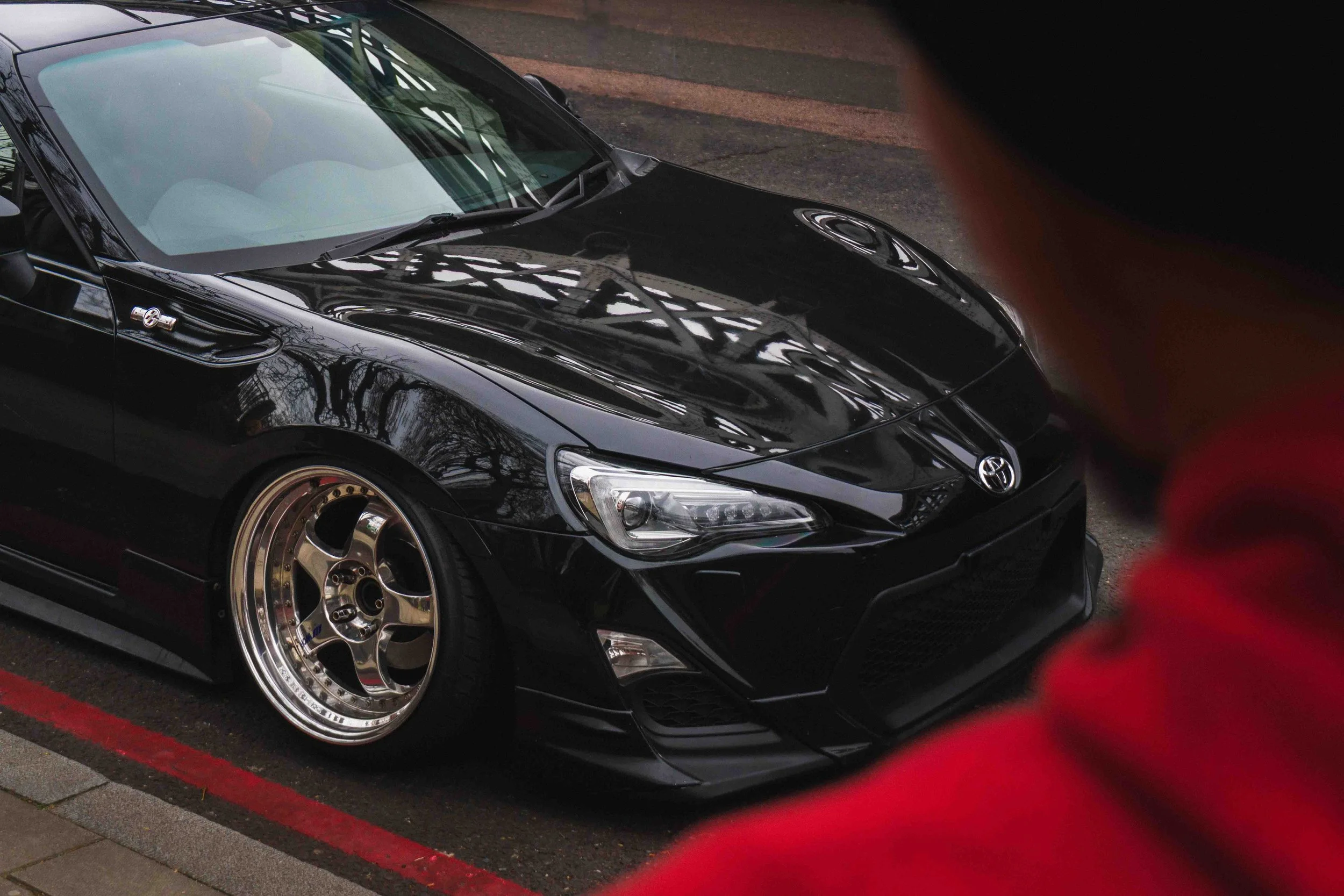 A black sports car with custom wheels parked near a curb, partially obscured by a person in a red hoodie.