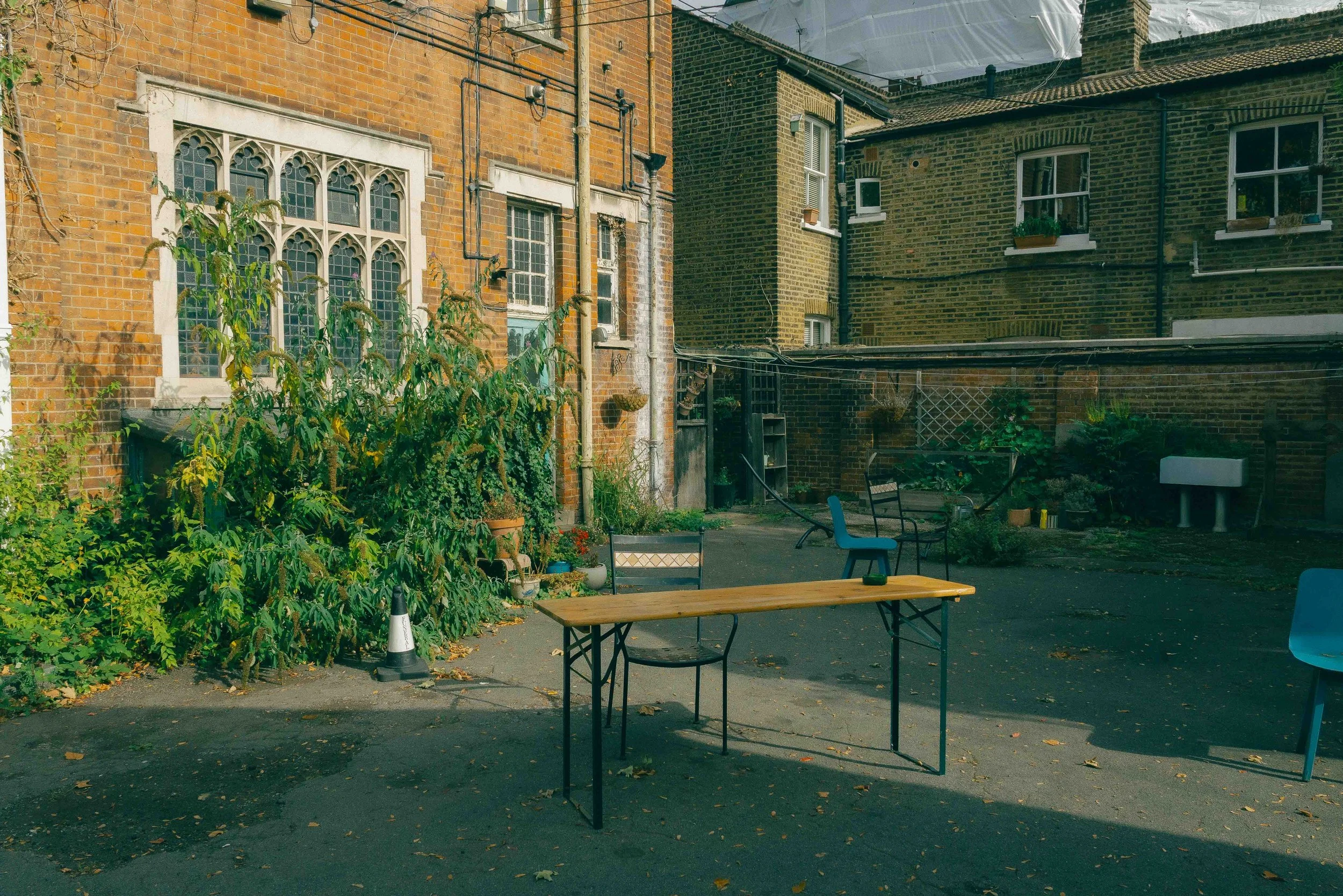 An outdoor courtyard with a wooden table, a few chairs, potted plants, and brick buildings in the background.