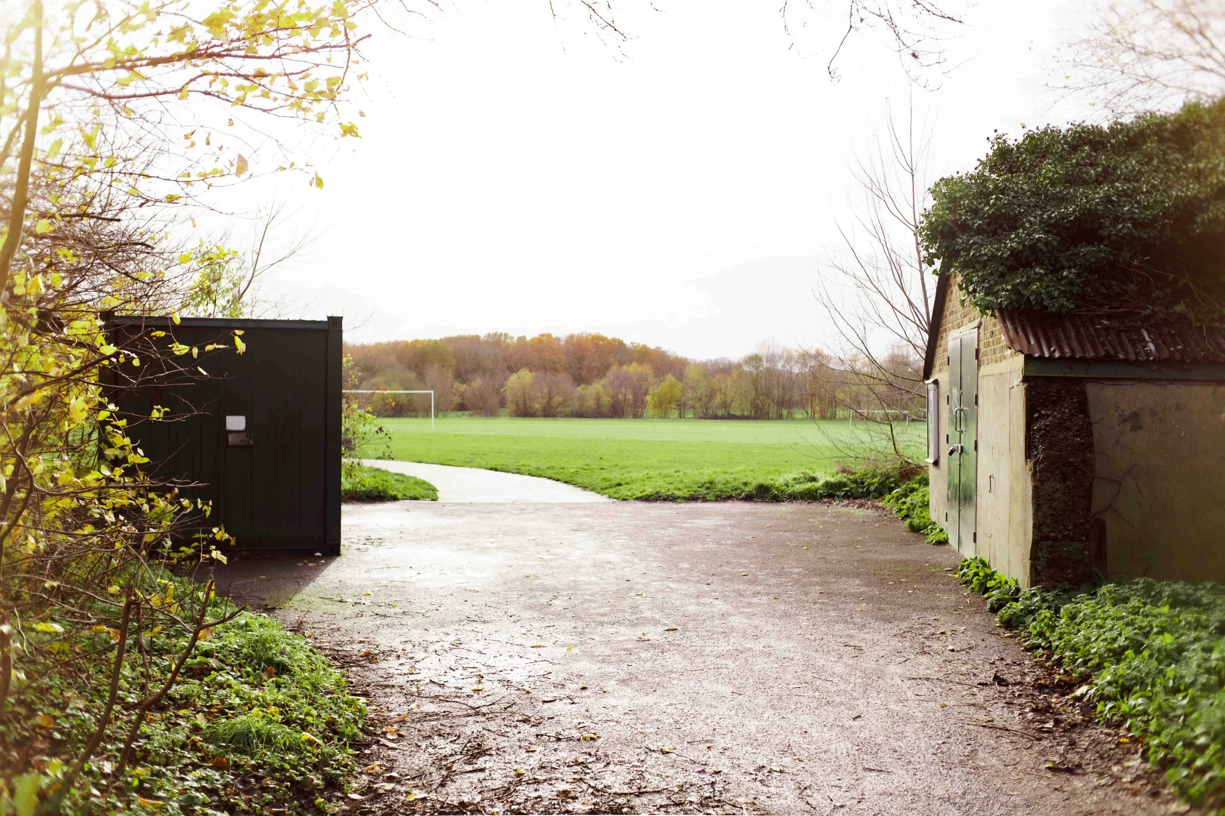 Outdoor scene featuring a dirt path leading to a grassy field with trees in the distance, flanked by two small sheds or structures on either side.