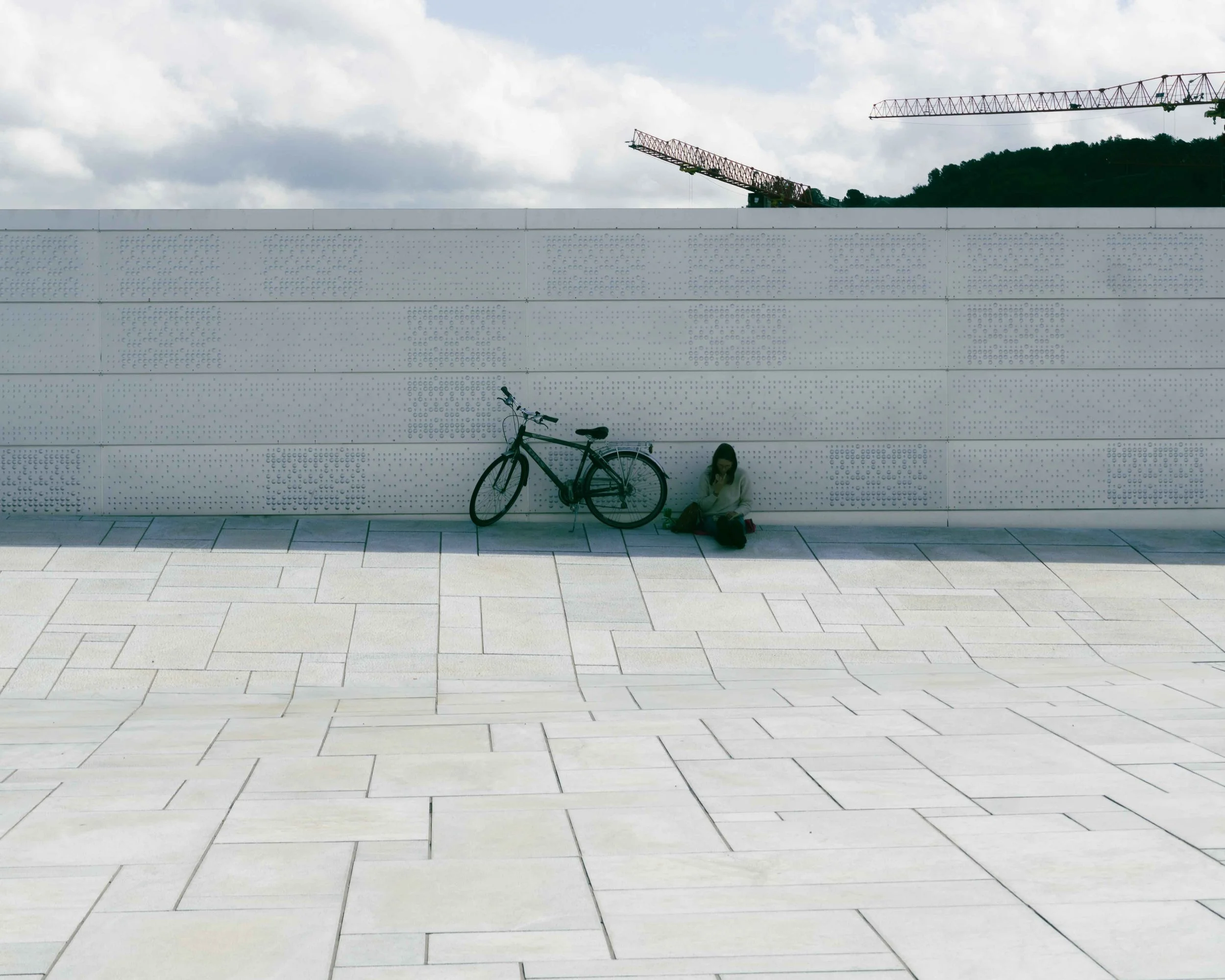 A woman sitting on the ground next to a bicycle, against a modern, white wall with a textured pattern, under a cloudy sky with two construction cranes in the background.