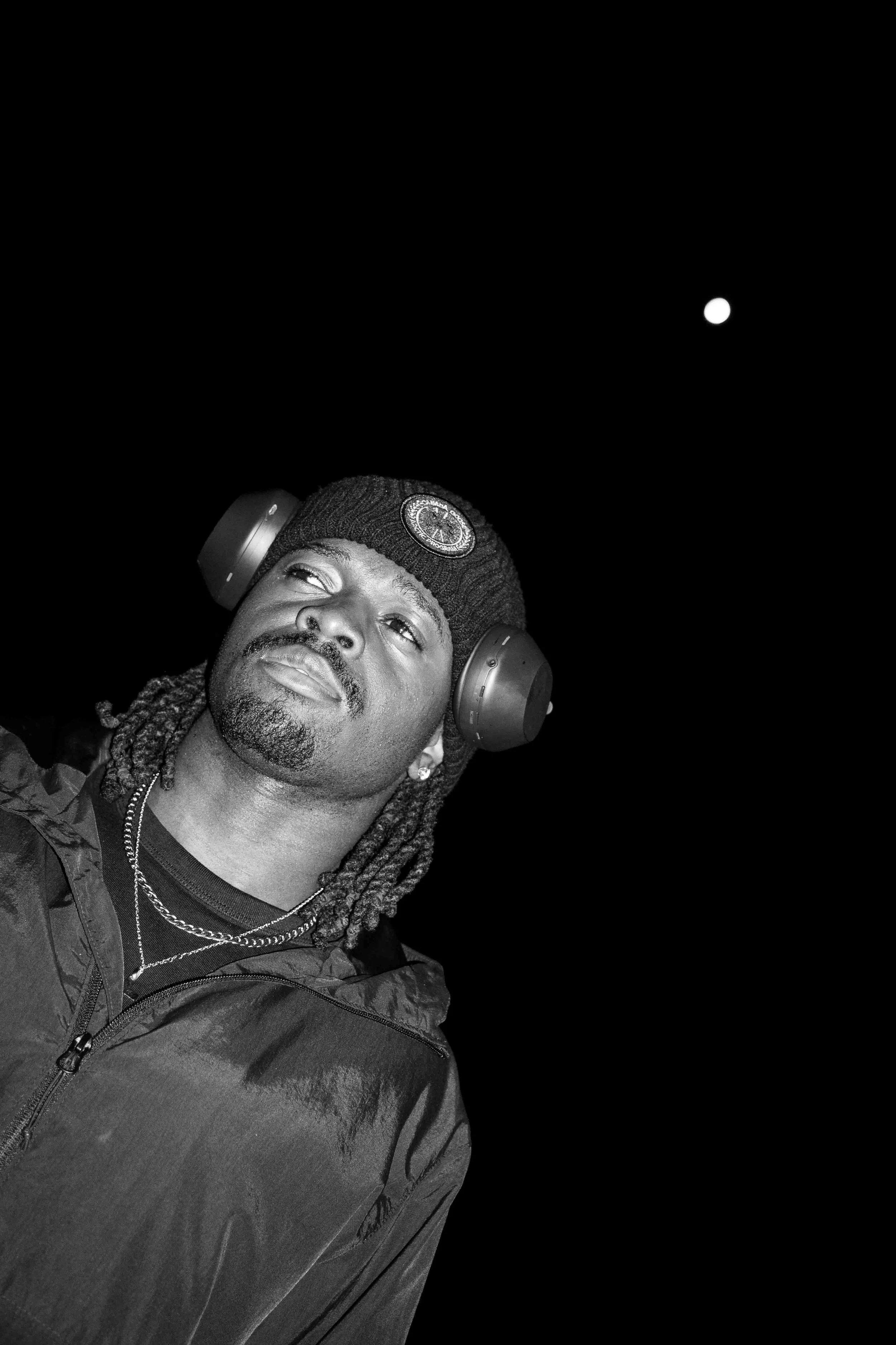 A man with dreadlocks and facial hair wearing headphones, a beanie with a circular patch, and a jacket, looking up against a night sky with a visible moon.