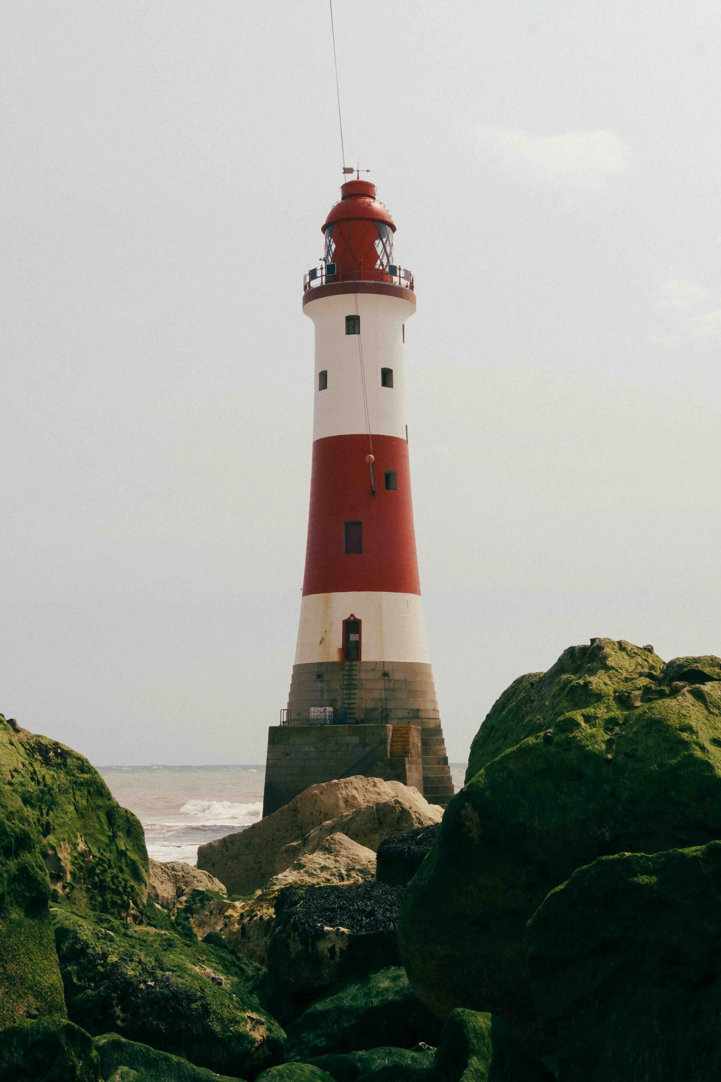 A tall white and red striped lighthouse on a rocky shoreline at the beach.