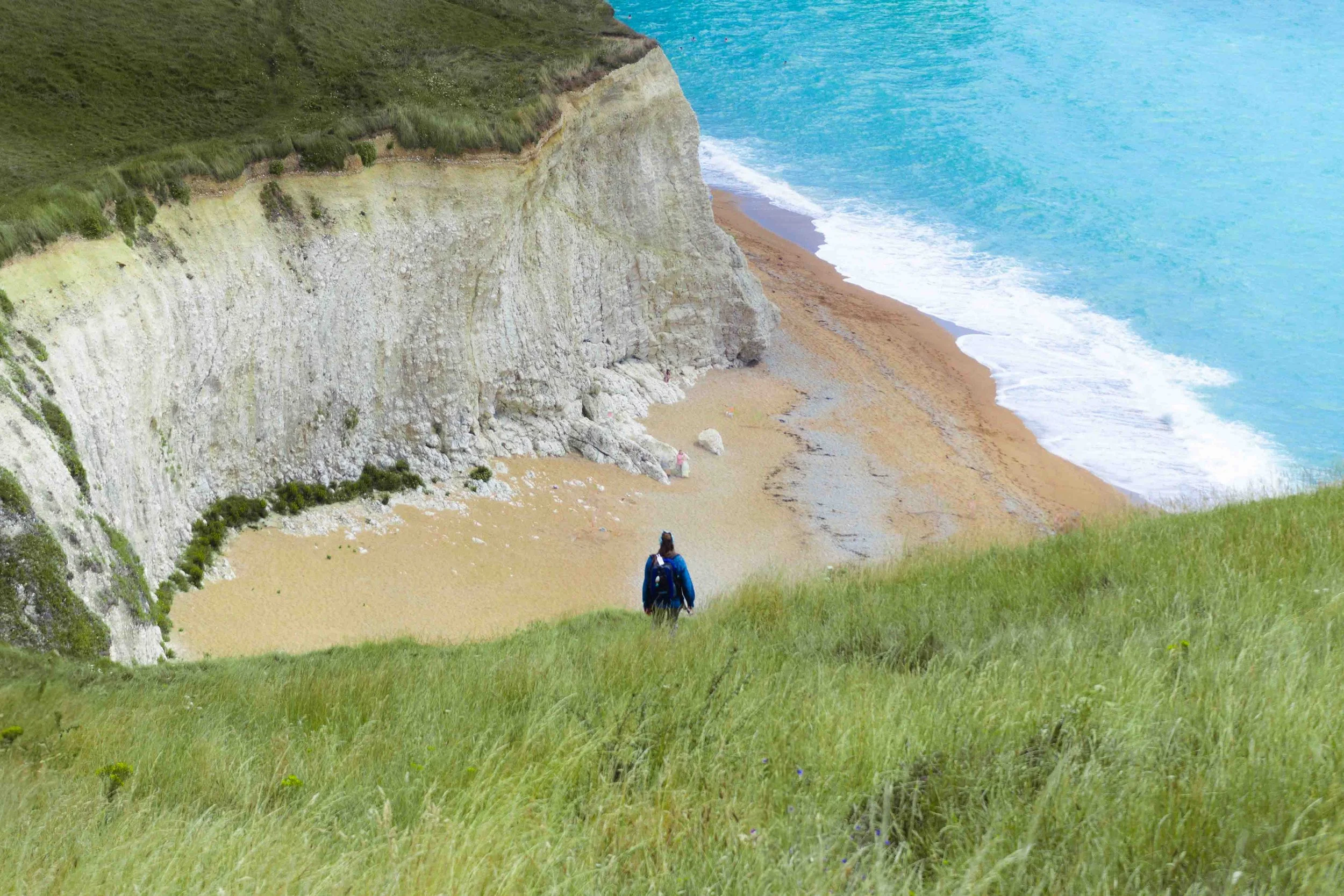 A person with a blue backpack walking down a grassy slope towards a beach with white chalk cliffs and ocean waves in the background.