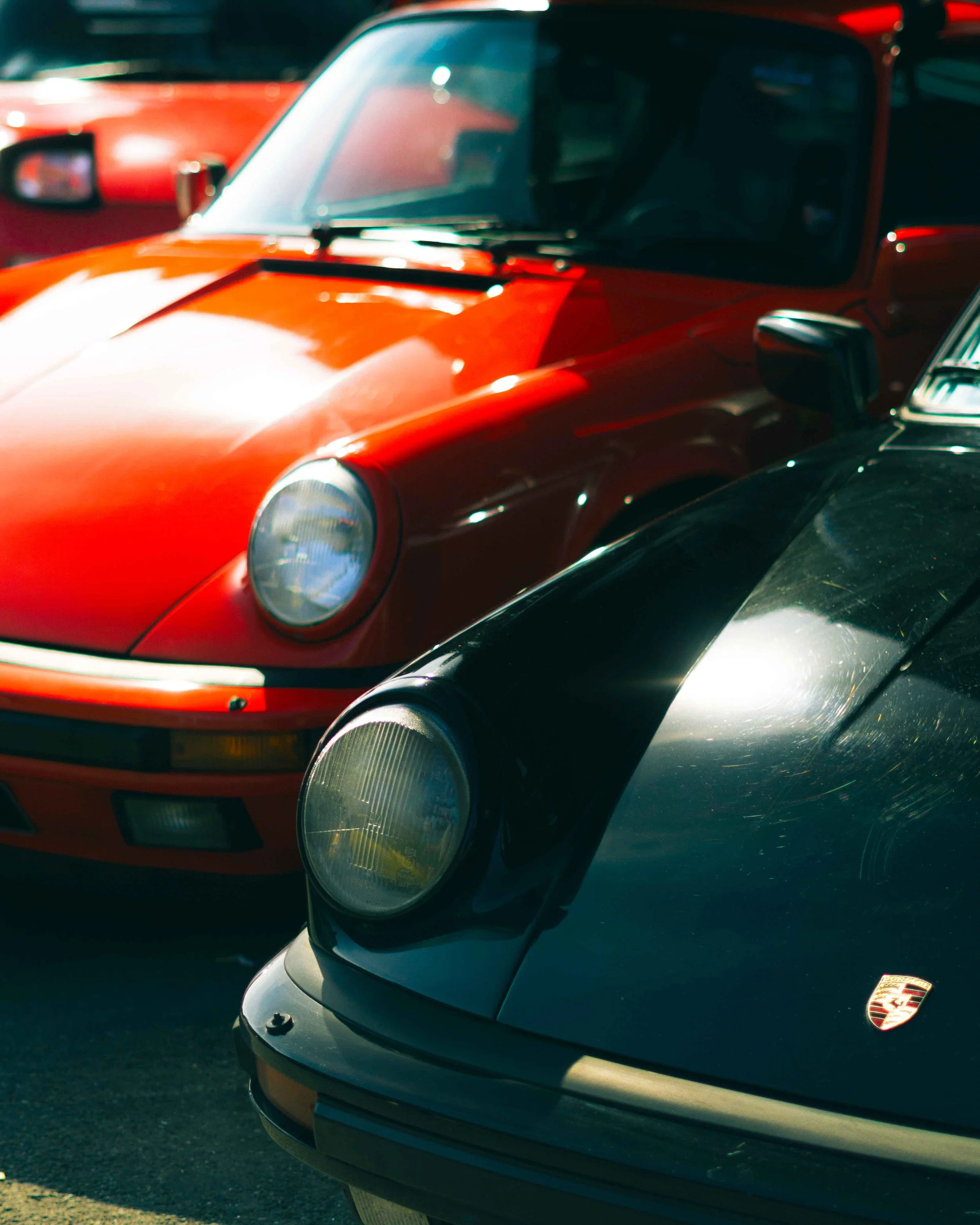 Close-up of two classic Porsche cars parked next to each other, one in black with a Porsche badge, and the other in bright red, both showing prominent front headlights.