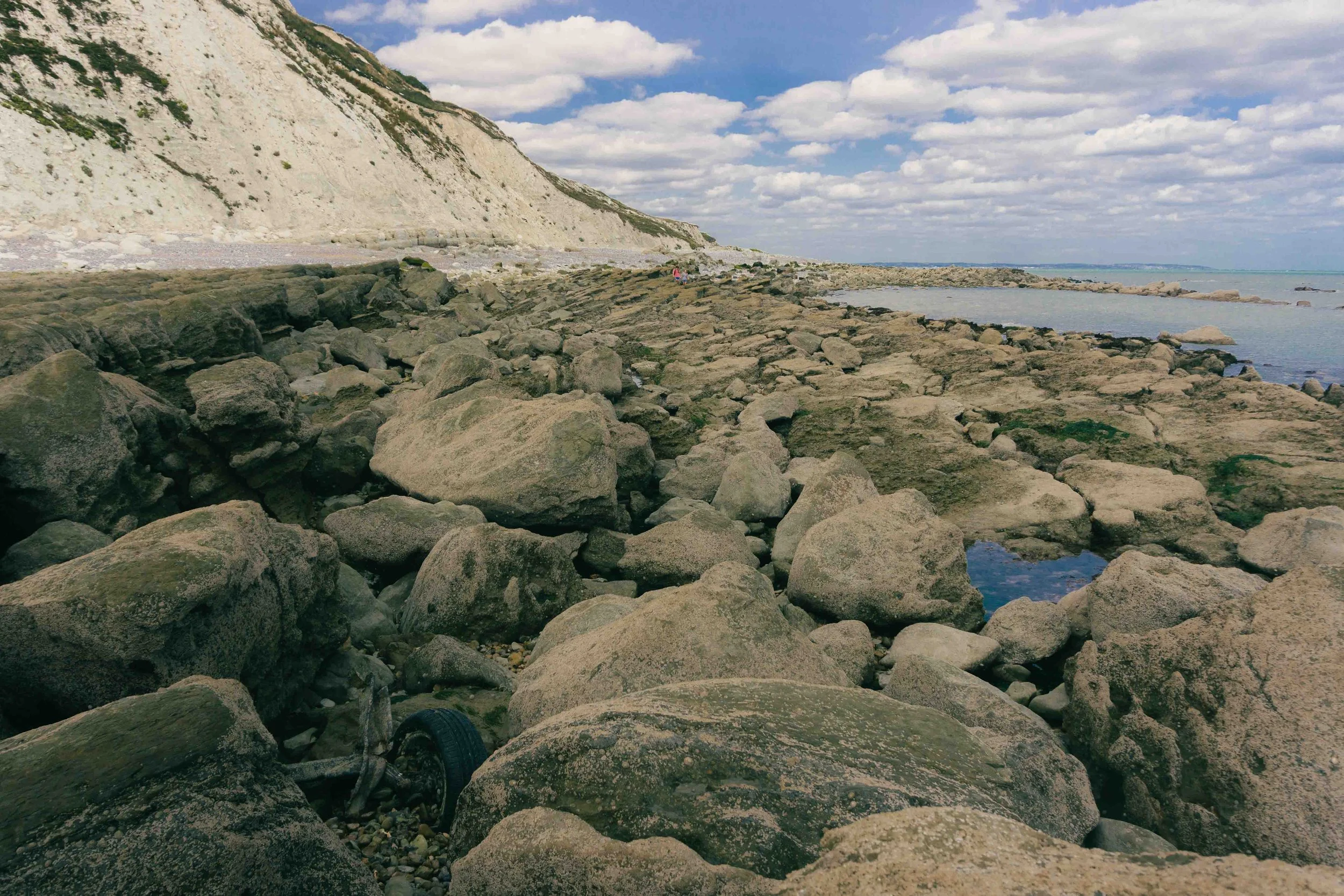 Rocky shoreline with large boulders, ocean, white chalk cliffs in the background, and cloudy sky.