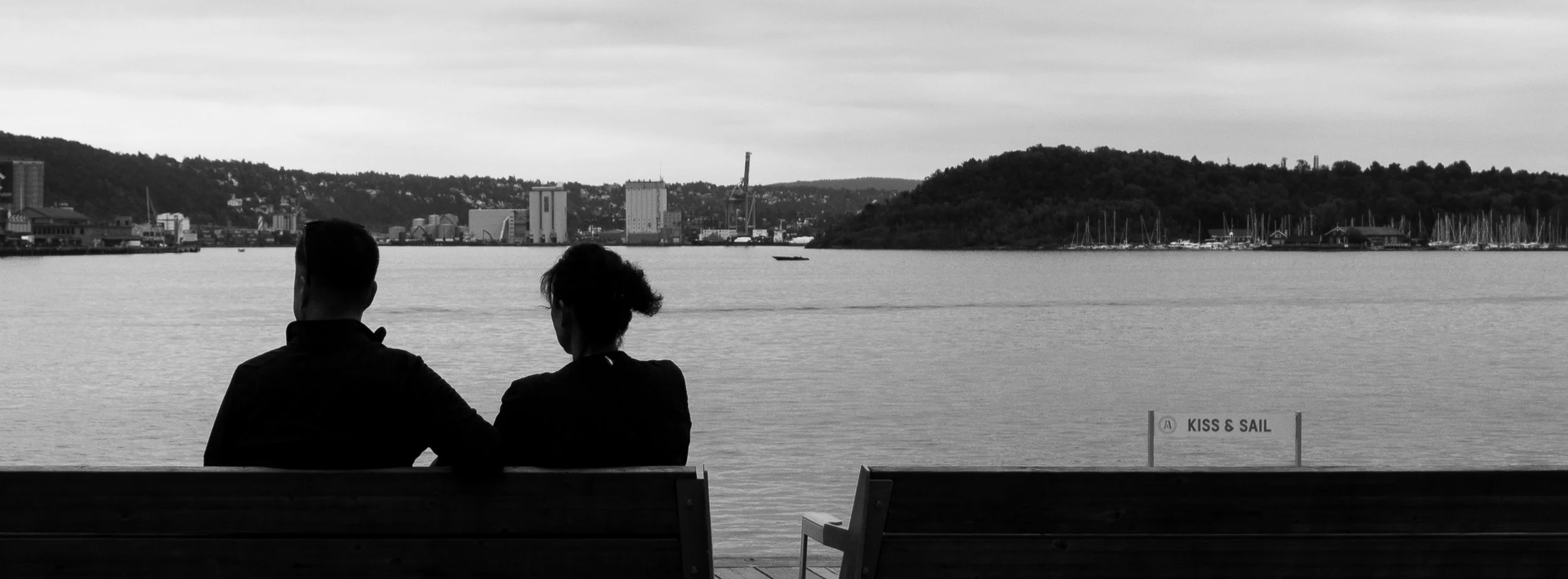 Two people sitting on a bench by the water, overlooking a city skyline and marina, in black and white.