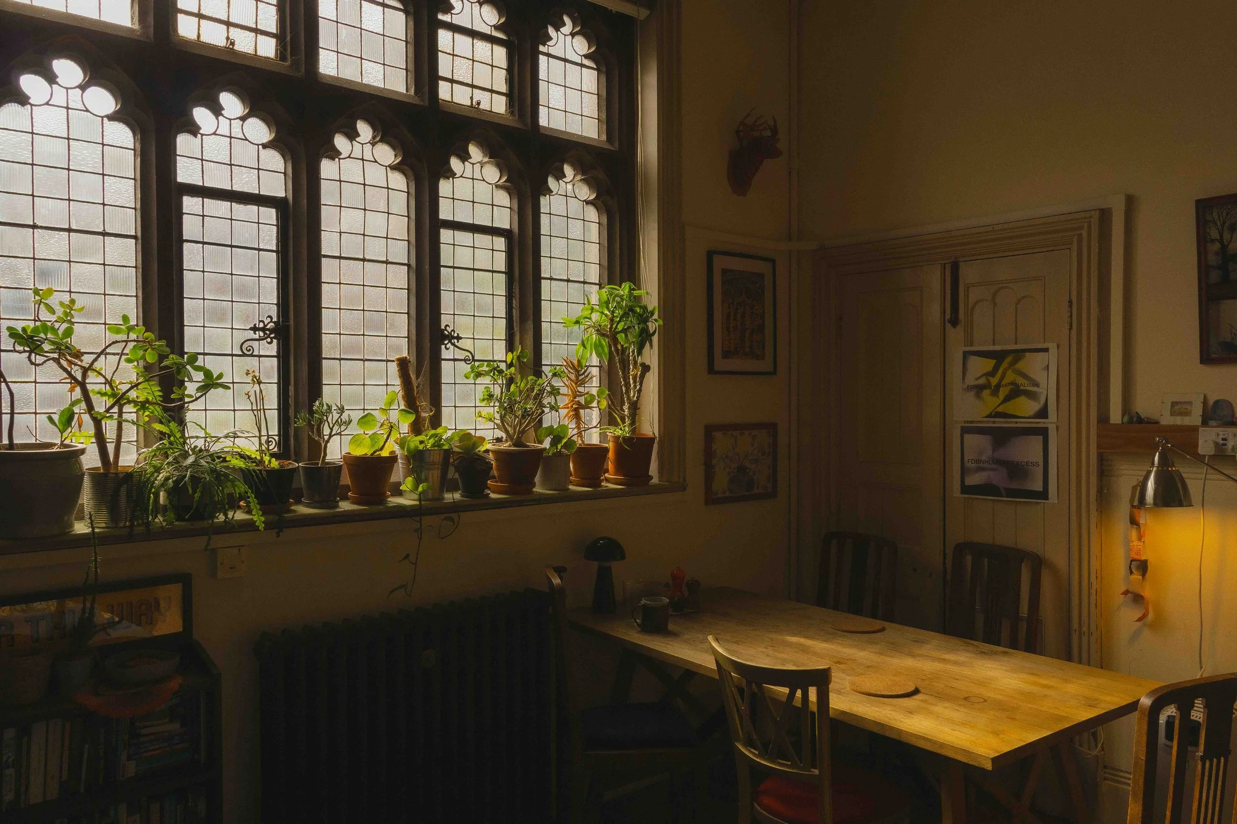 An interior room featuring large arched windows with multiple glass panes, adorned with a row of potted plants on the windowsill. The room contains a wooden dining table with chairs, and is decorated with framed artwork on the walls and a mounted dee