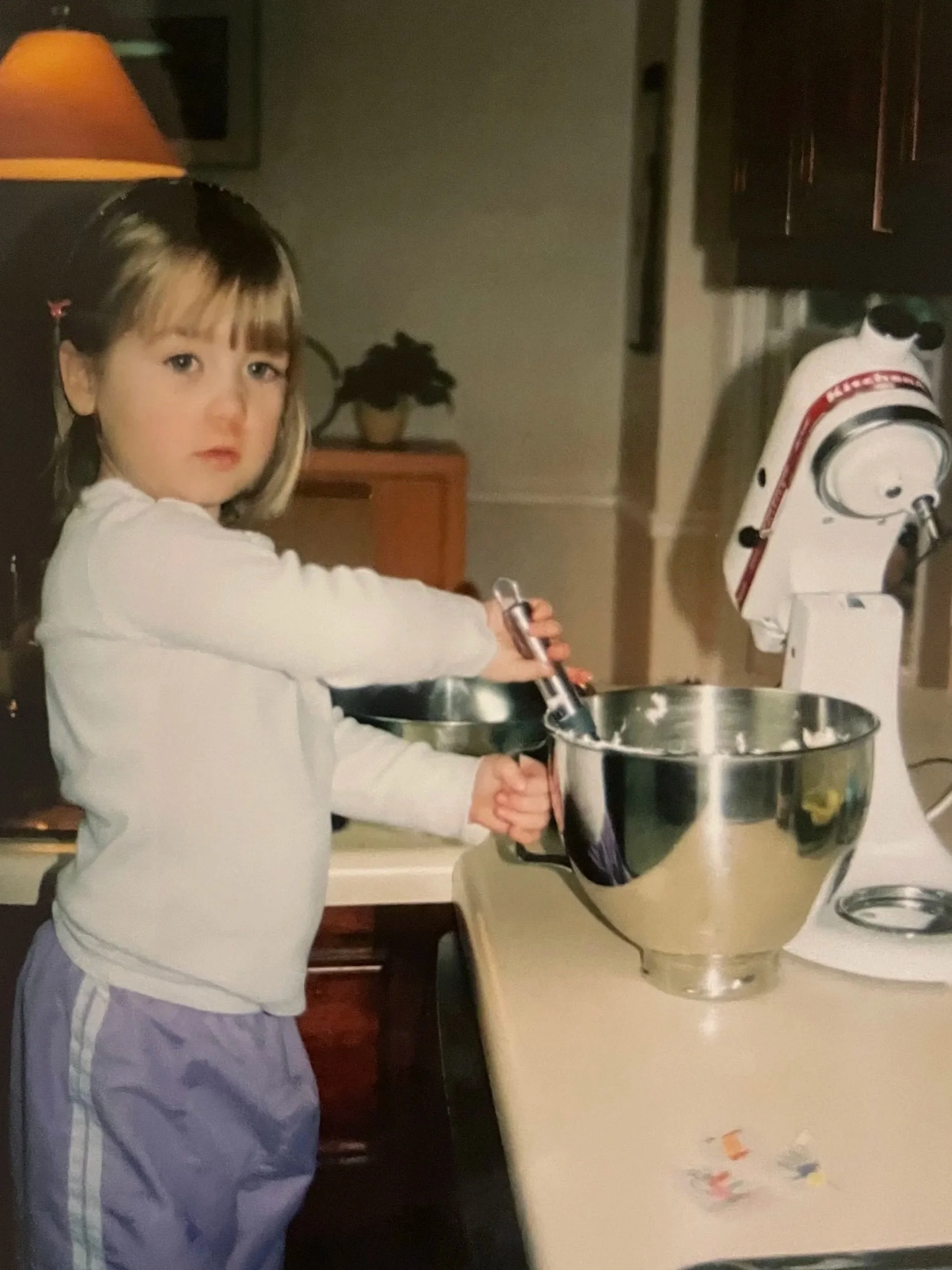 Young girl with brown hair and bangs stirring a mixture in a large metal mixing bowl in a kitchen.