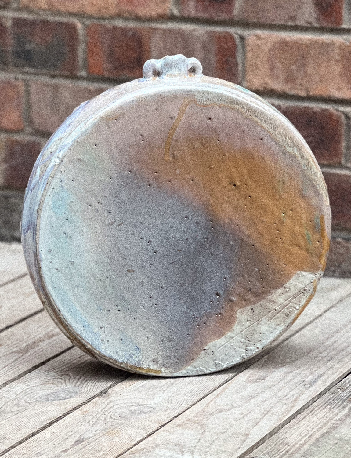 Close-up of a ceramic vase or container, showing its textured interior and exterior with a rustic, glazed finish, placed on a wooden surface against a brick wall background.