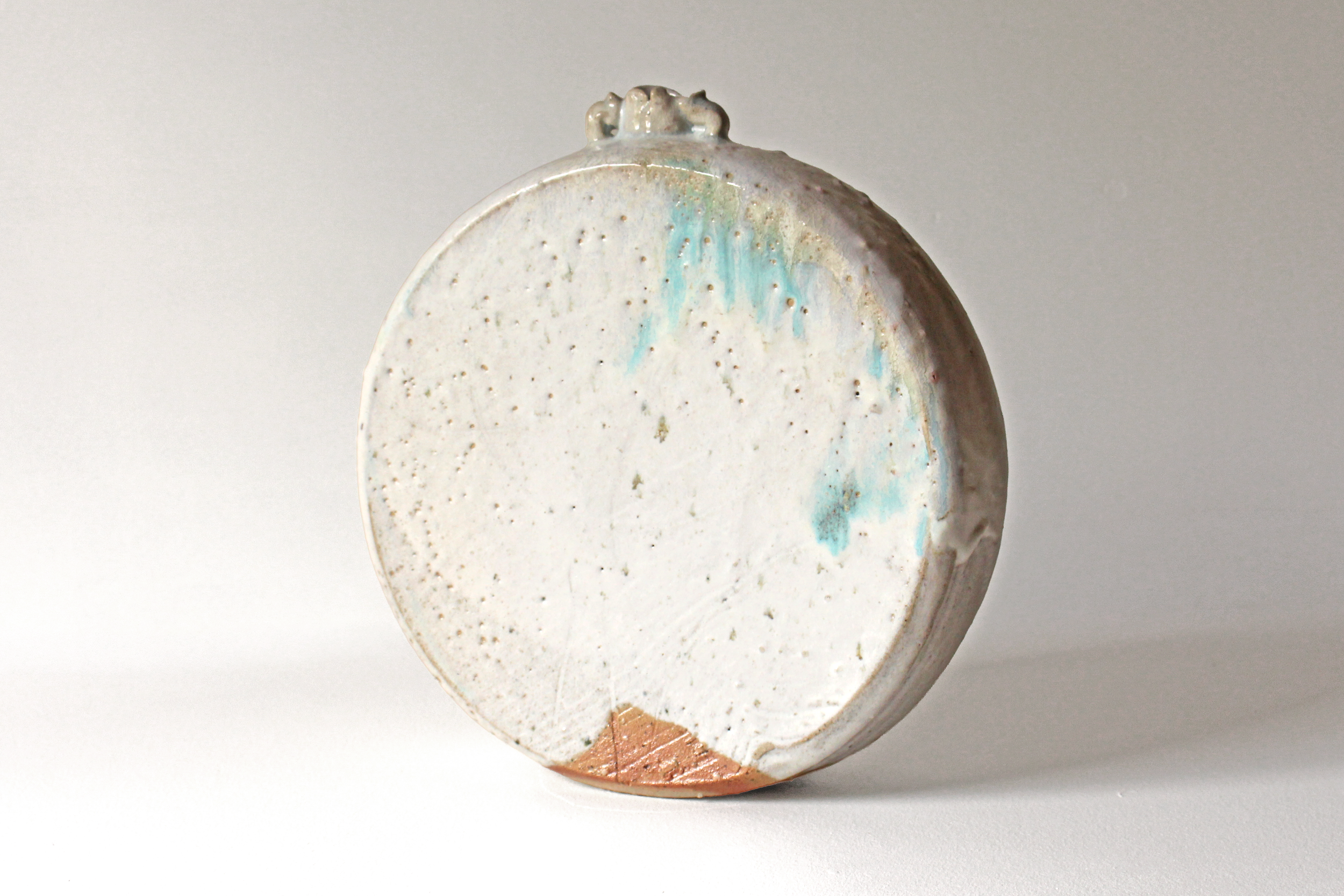A ceramic pottery piece with a rounded, flat base and a textured, uneven surface, decorated with subtle blue, green, and brown glaze accents.