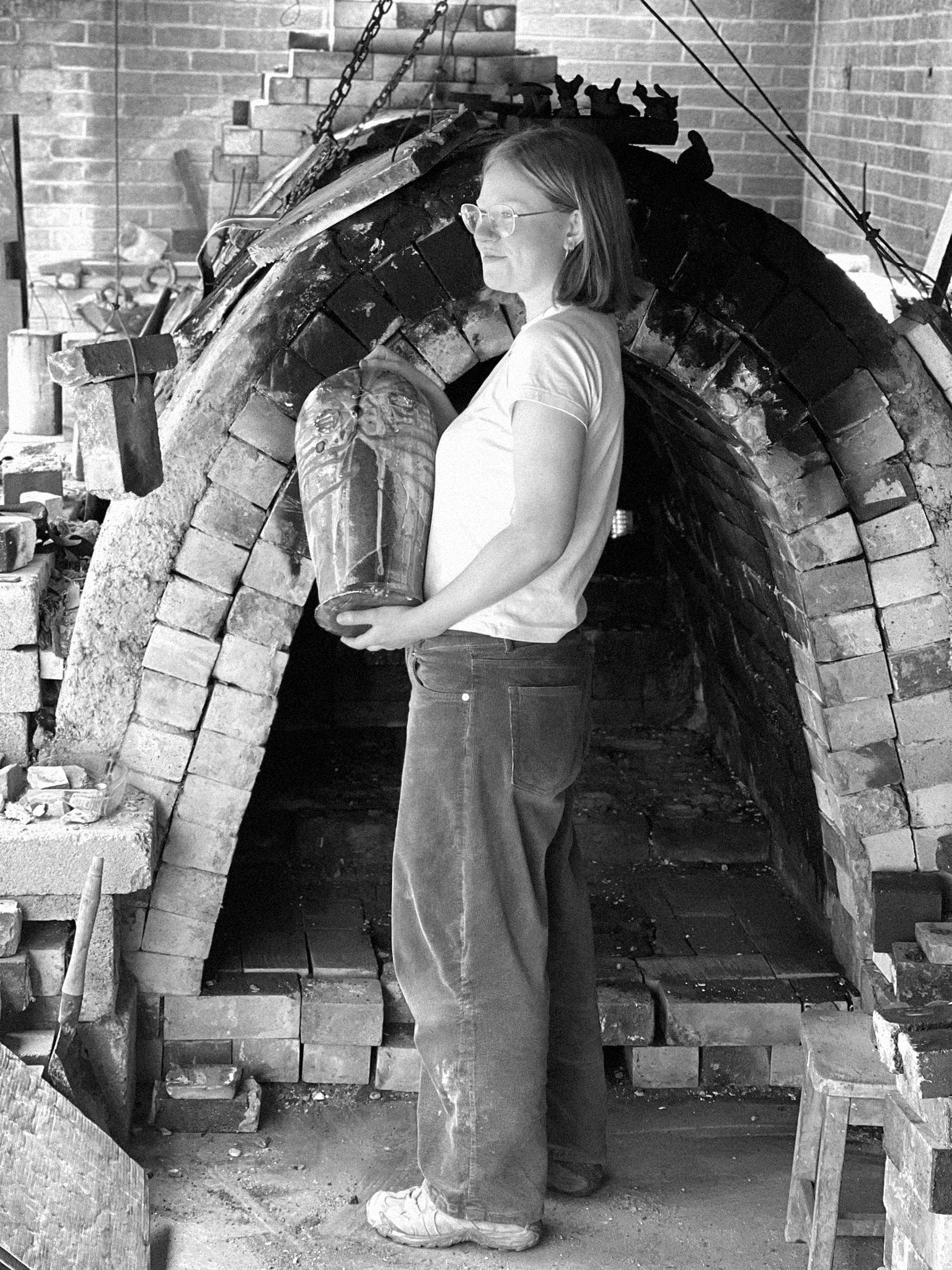 A young woman with shoulder-length hair and glasses is standing near a wood kiln, holding a large jar and looking to her left.