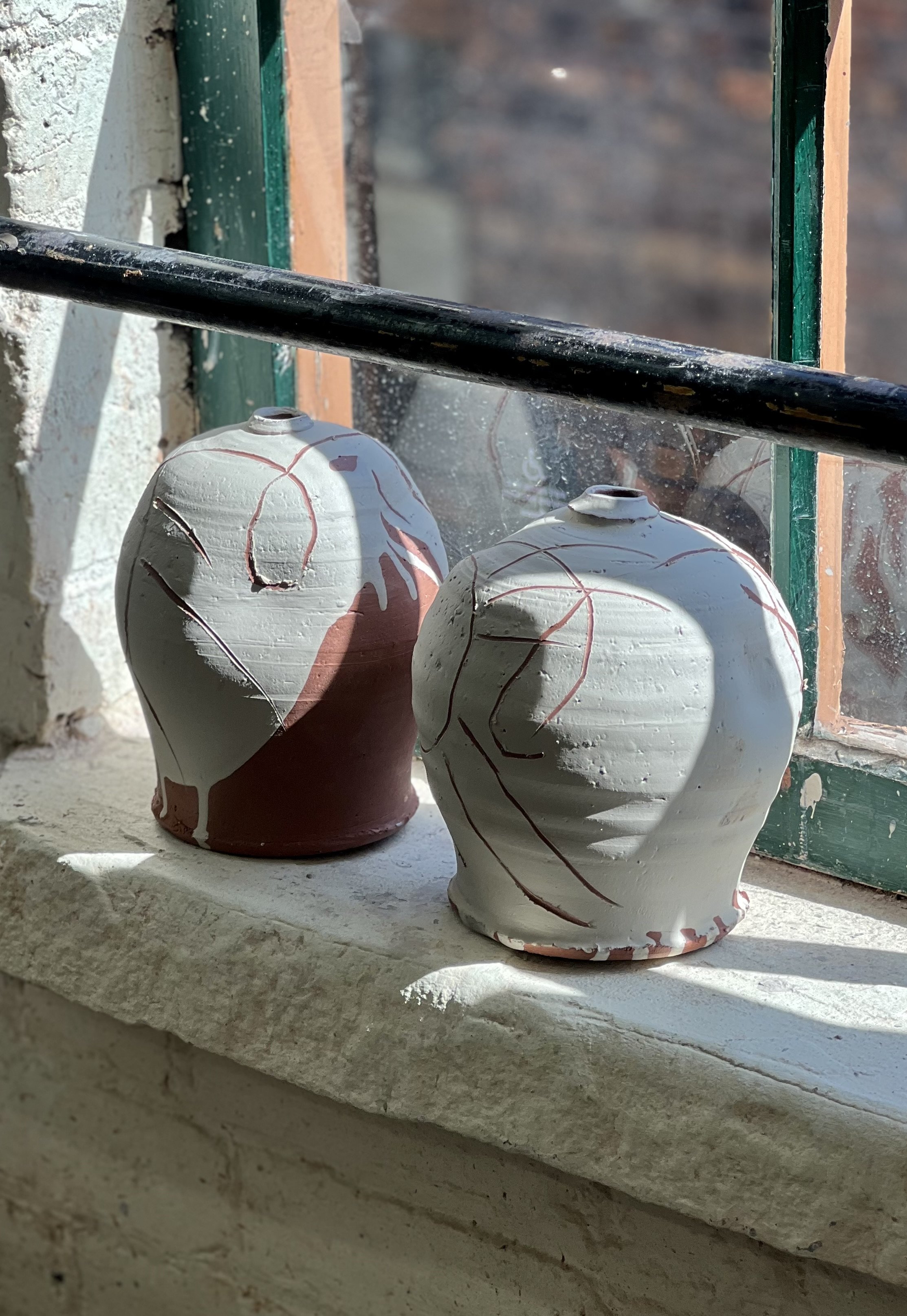 Two ceramic vases with abstract line designs on a windowsill in sunlight, with a green window frame and a brick wall visible outside.