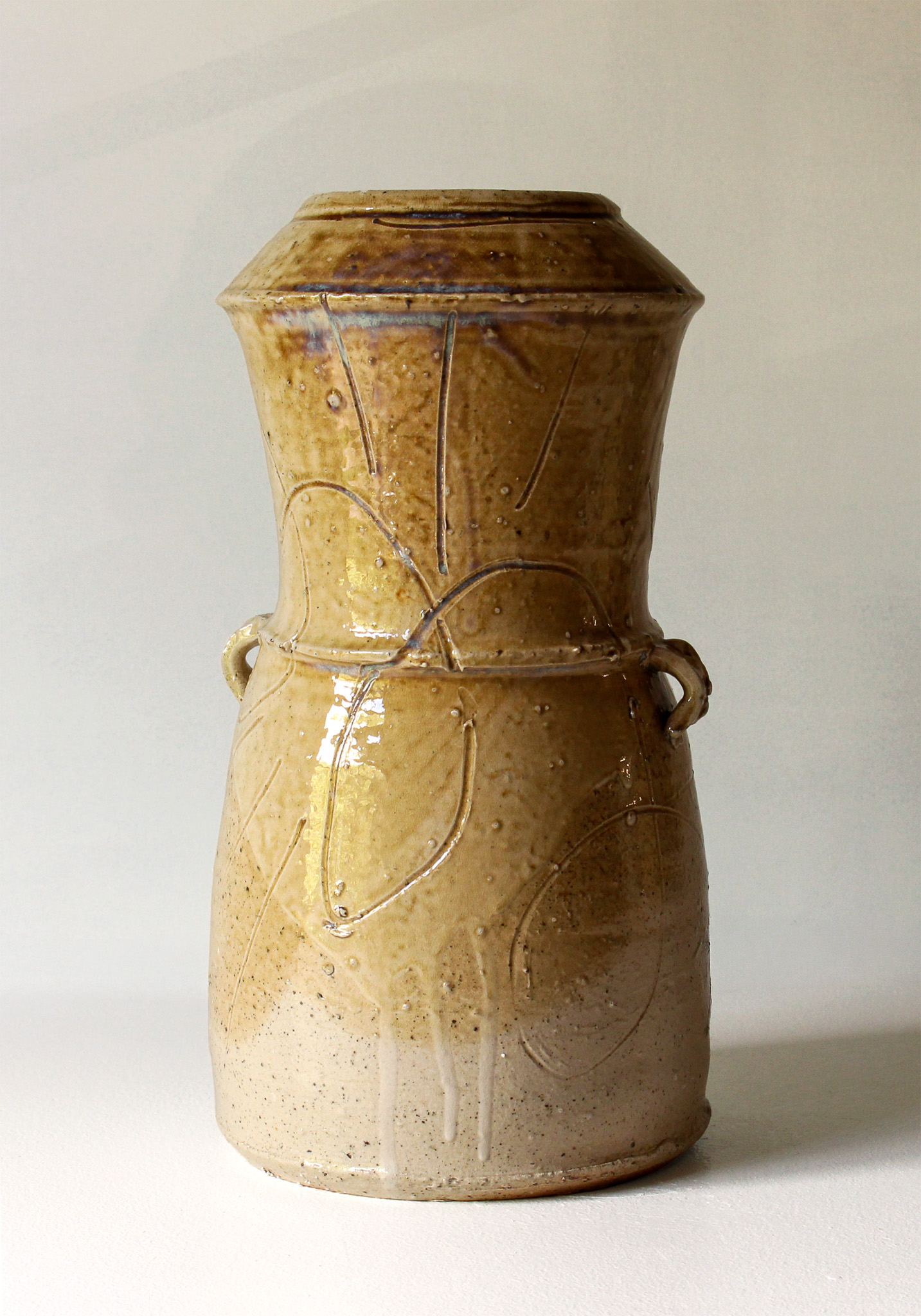 A ceramic vase with a narrow top and handles on the sides, glazed in earthy tones of brown and beige, decorated with carved line patterns and small bubbling dots, set against a plain, light-colored background.