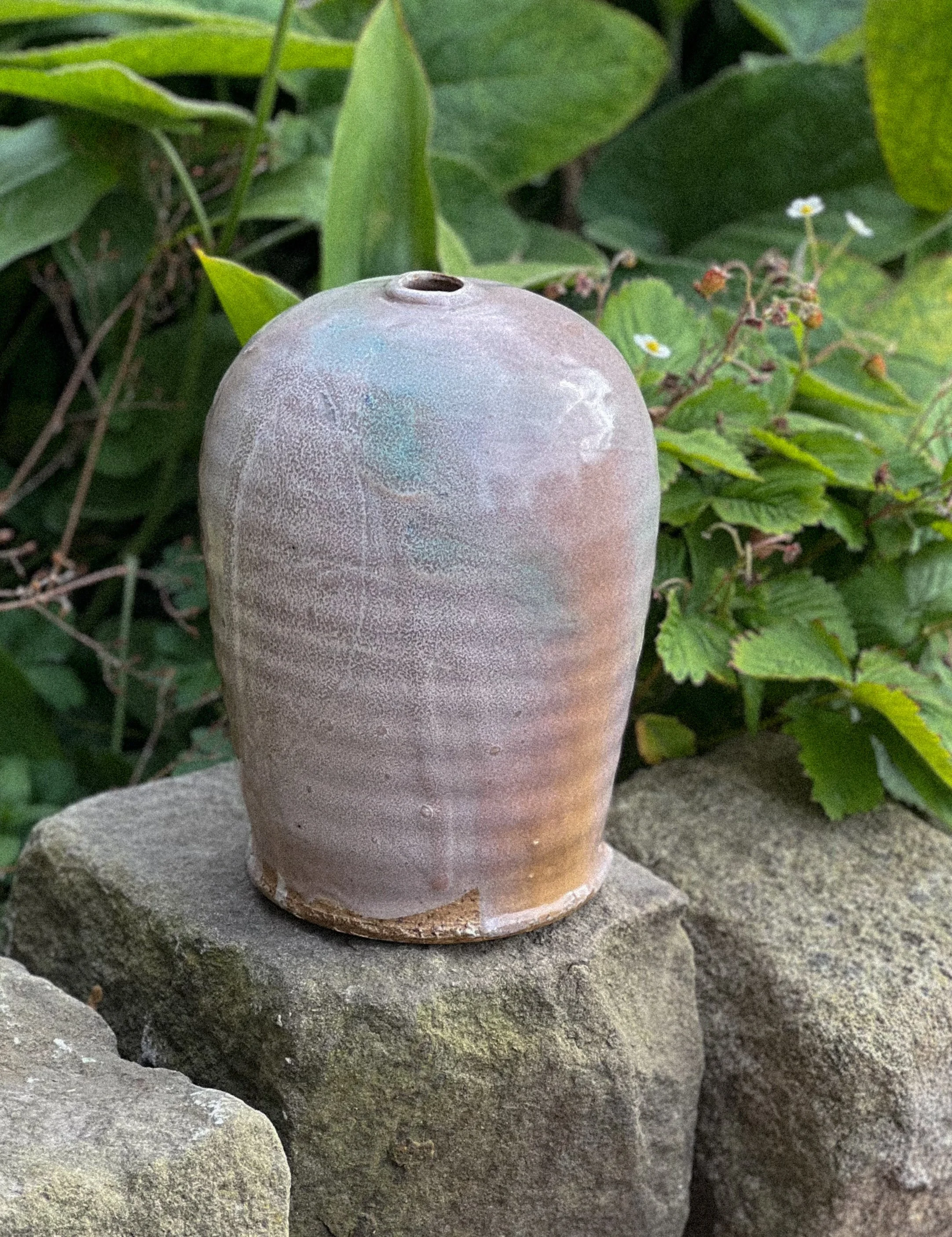 A ceramic vase with a rounded top and textured surface, positioned on a large stone, surrounded by green plants and small white flowers.