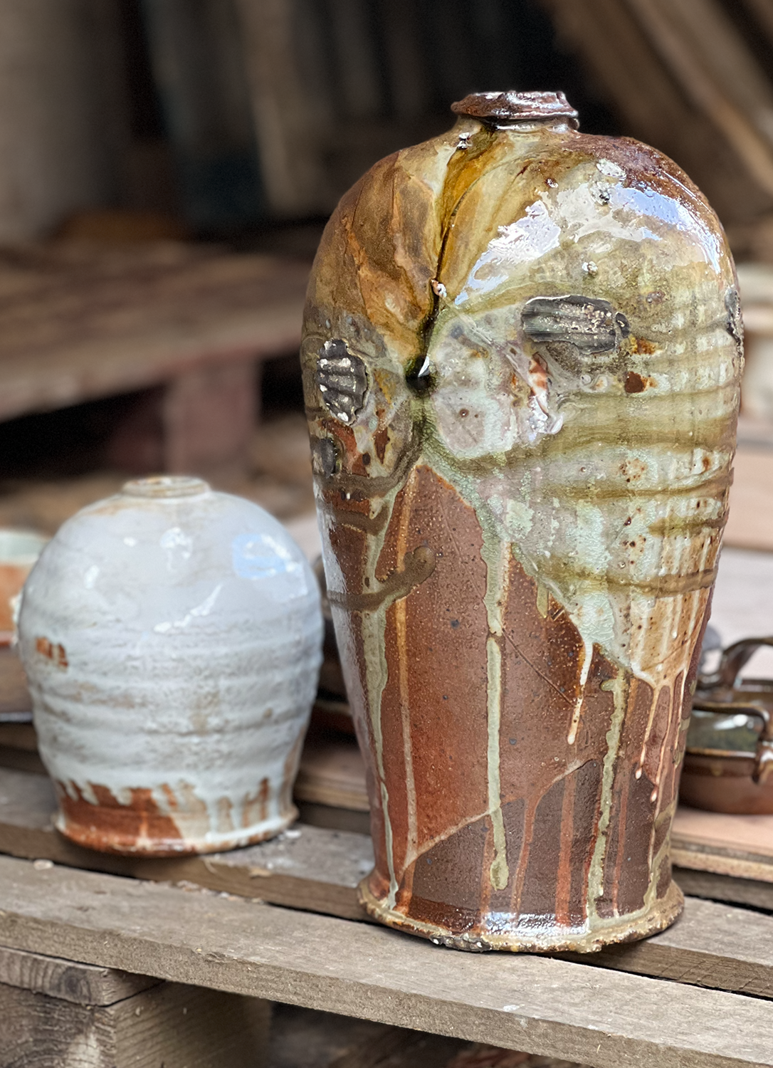 Close-up of two ceramic vases on a wooden surface, with a rustic background. The larger vase has a textured, multicolored glaze with dripping effects, predominantly in shades of brown, cream, and orange, while the smaller vase is white with a similar