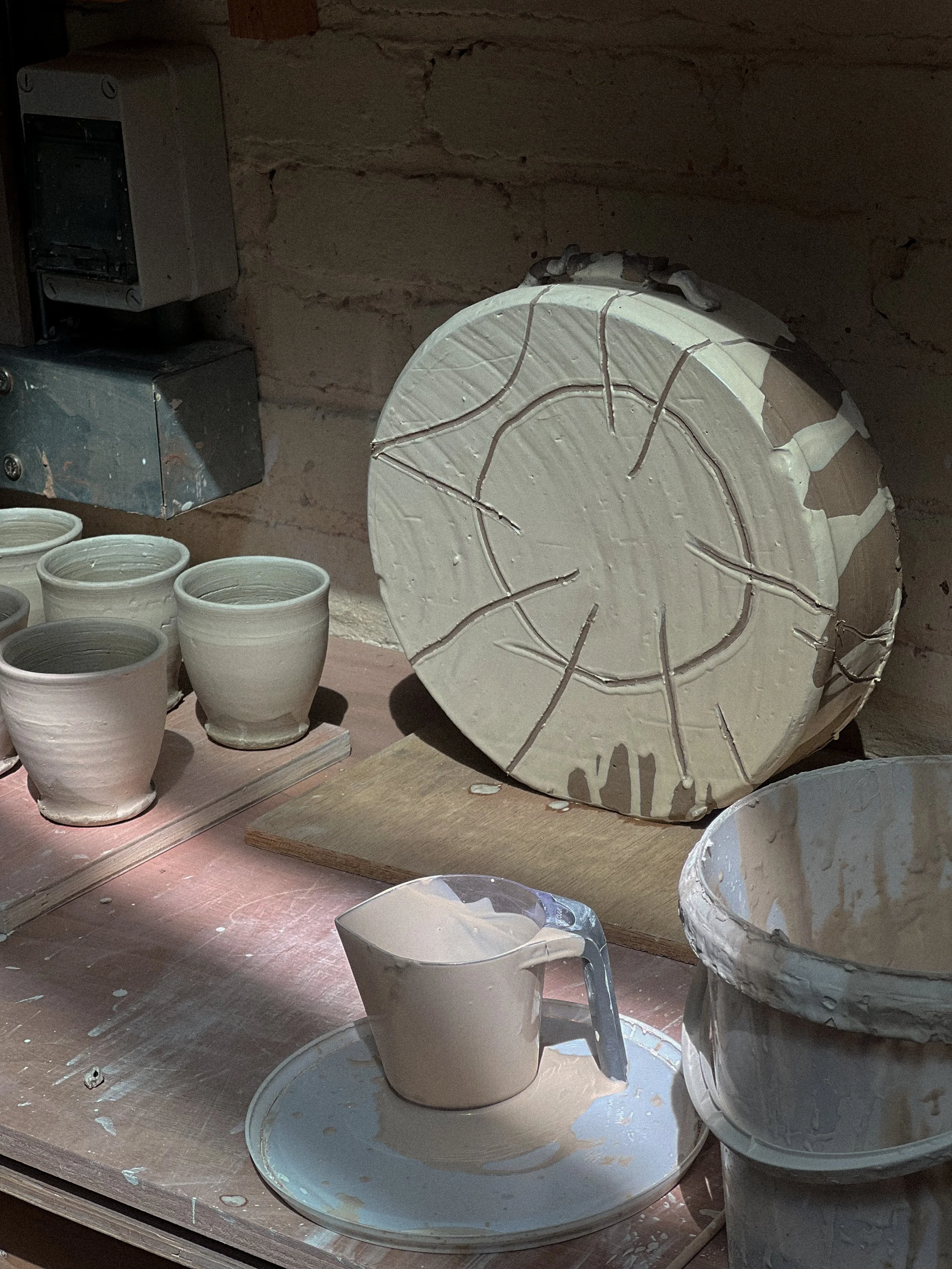 Pottery workspace with various unfinished ceramic pots and bowls, some on a wooden workbench, with a wheel and tools for shaping clay.