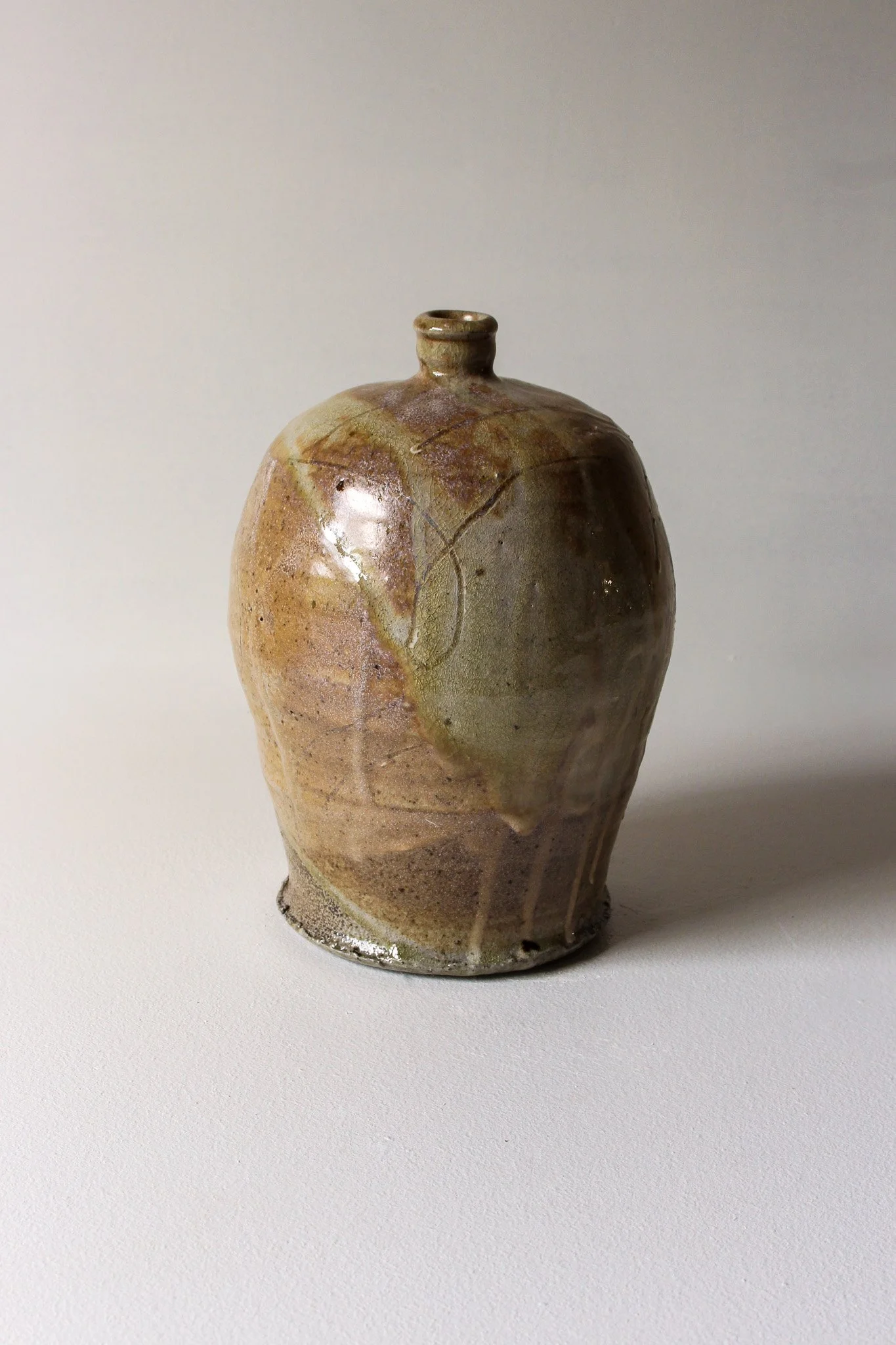 A ceramic vase with a narrow neck, decorated with earthy tones and crackled glaze, sitting on a white surface against a plain light background.