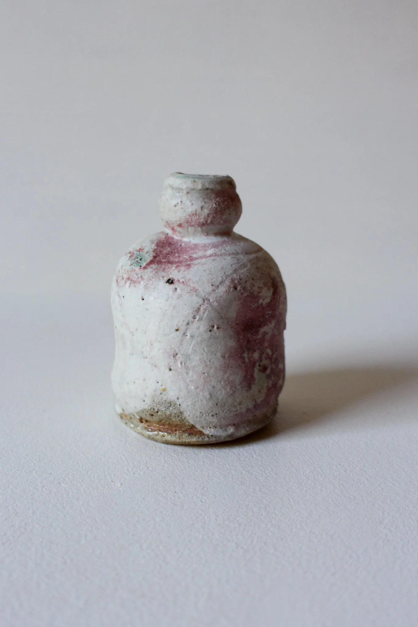 Small handmade ceramic vase with a rounded body, narrow neck, and a wide opening, glazed in blue with reddish accents and a textured surface.