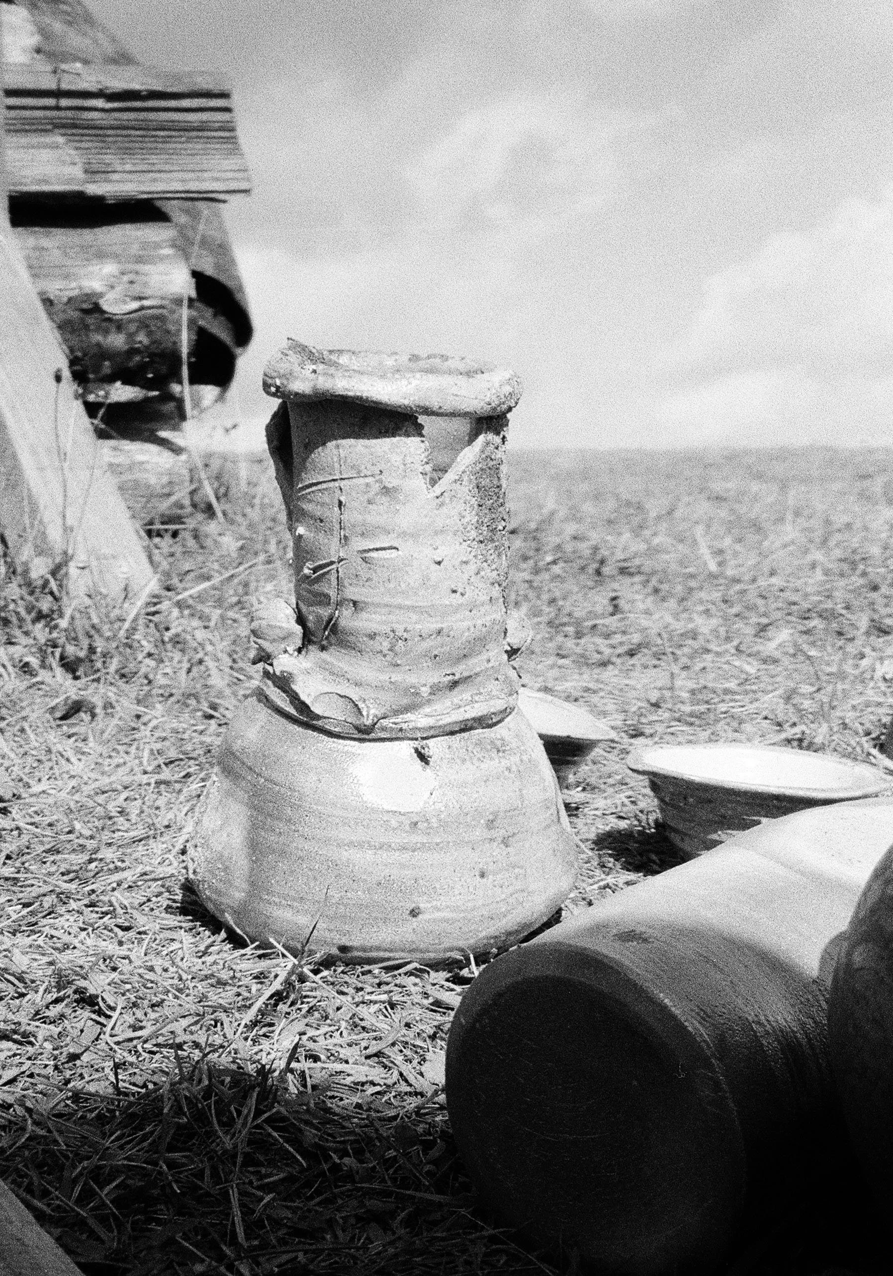 Black and white photo of a ceramic vase, placed outdoors on dry grass, with a cloudy sky in the background.