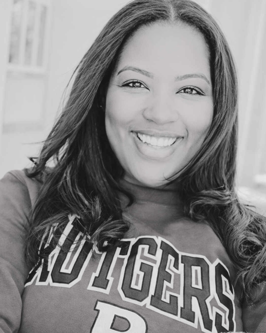 Black and white photo of a smiling woman with long wavy hair, wearing a sweatshirt with the word 'Rutgers' on it. Dr. Beauty