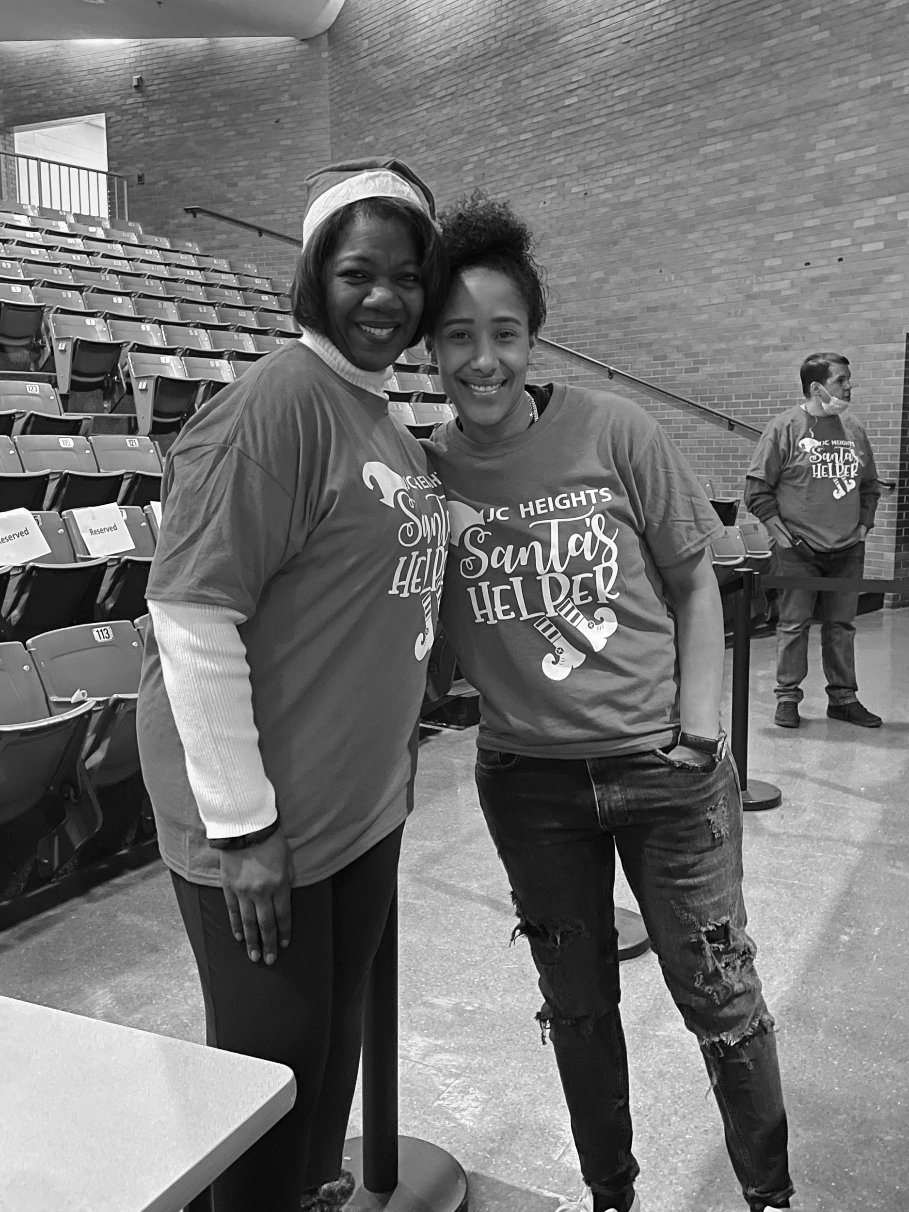 Two women posing together indoors, both wearing 'JC Heights Santa Helper' T-shirts, one is wearing a Christmas hat, and a third person in the background is also wearing a similar T-shirt.
