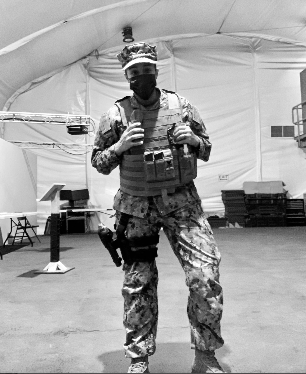 A soldier in camouflage uniform stands inside a large tent, wearing a tactical vest, a face mask, and a cap, with equipment attached to his belt.