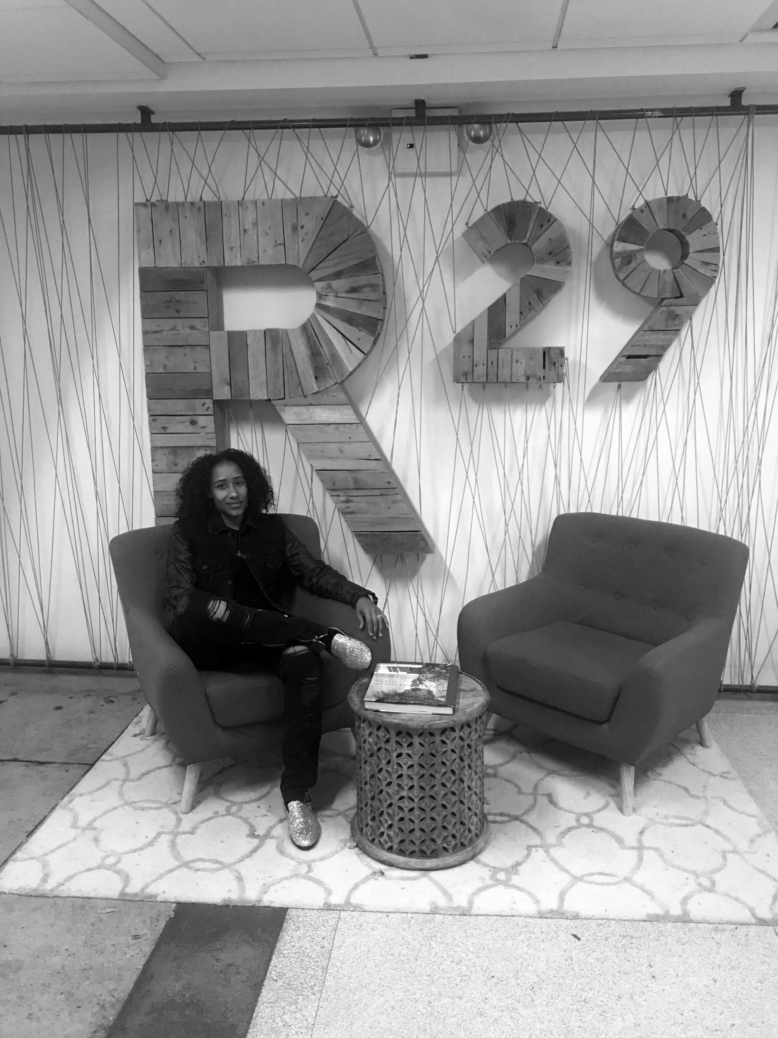 A woman sitting on a chair with two large wooden numbers 'R2' in the background, decorated with wooden planks, in a modern indoor setting.