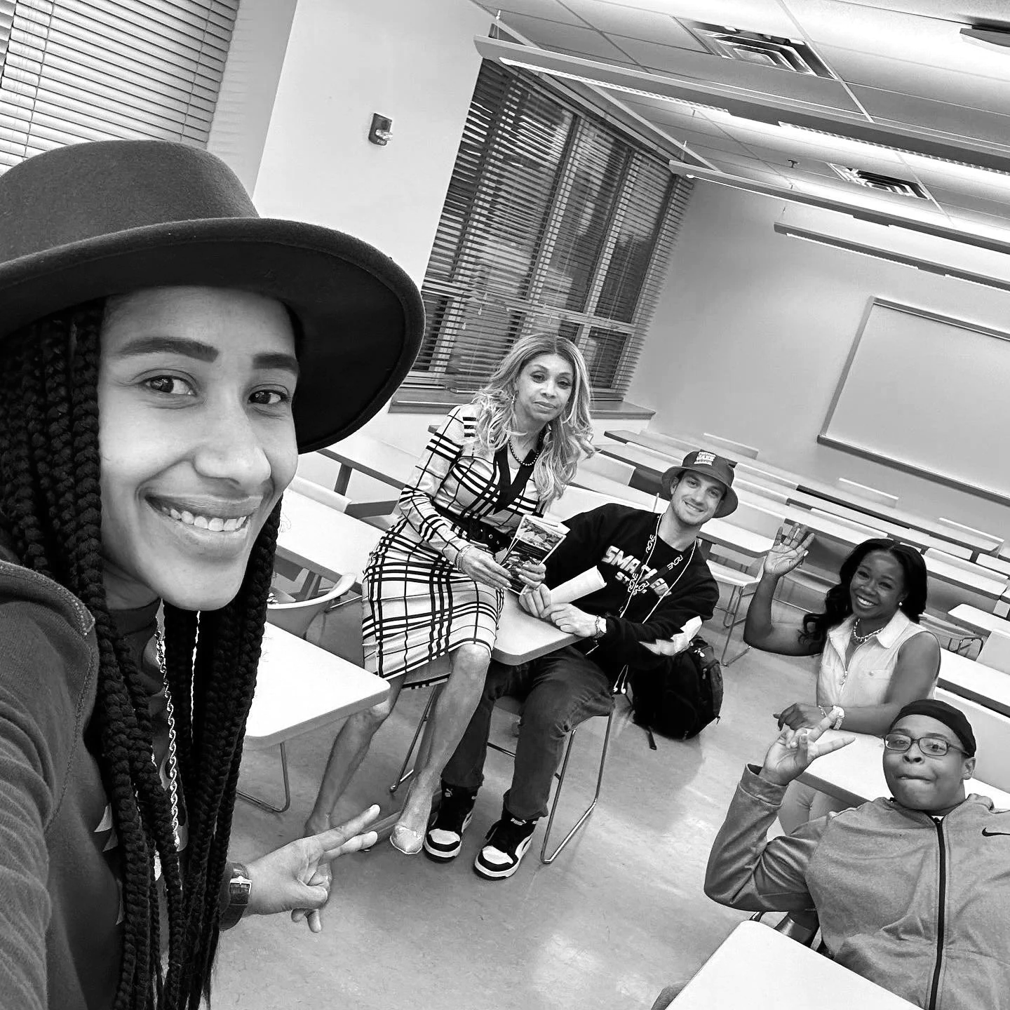 Five diverse people sitting and standing in a classroom, smiling and waving at the camera. The woman in the foreground is taking a selfie, wearing a large hat and braided hair.