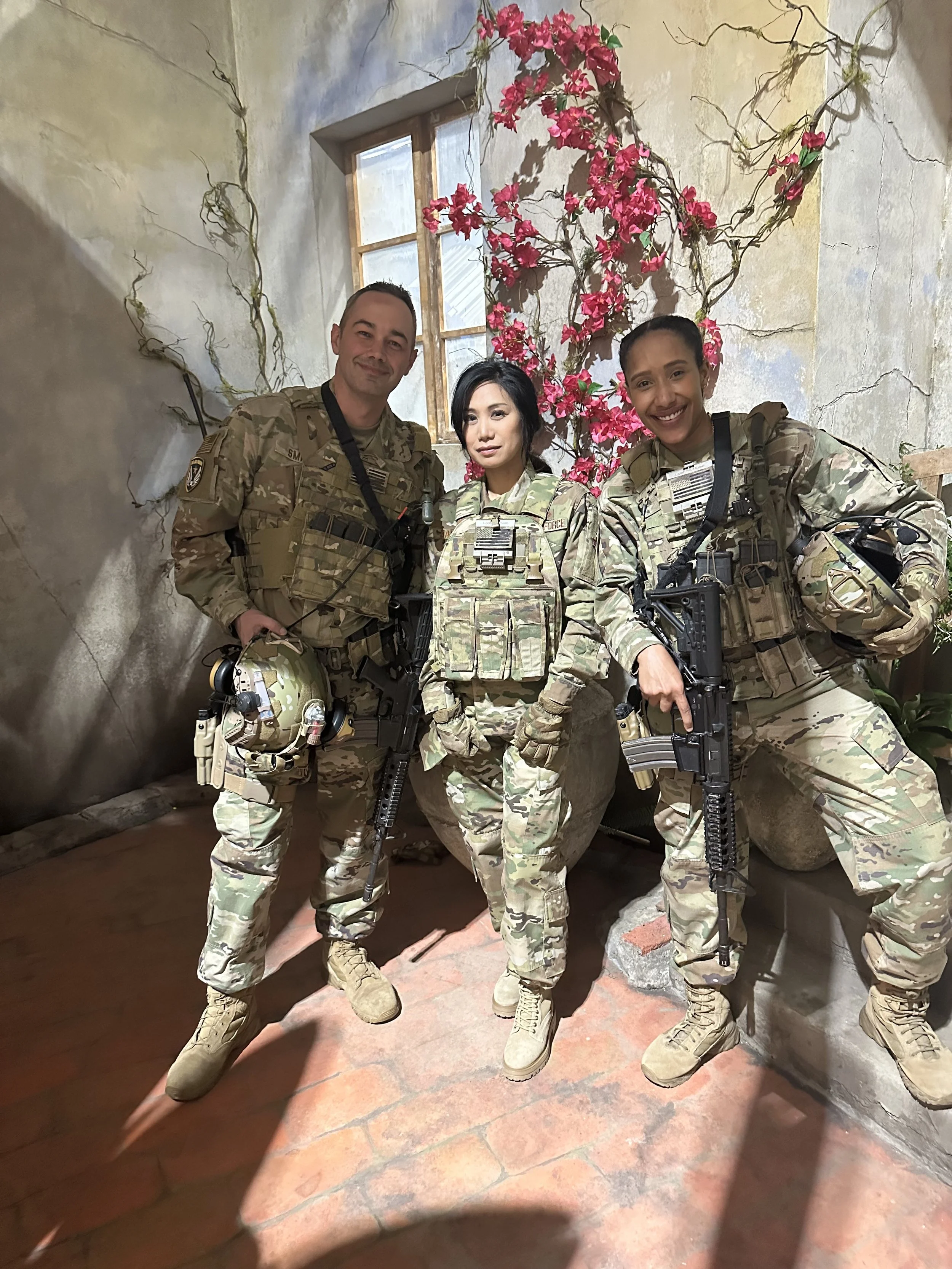 Three soldiers in camouflage uniforms posing inside a building with pink flowers and a window in the background.