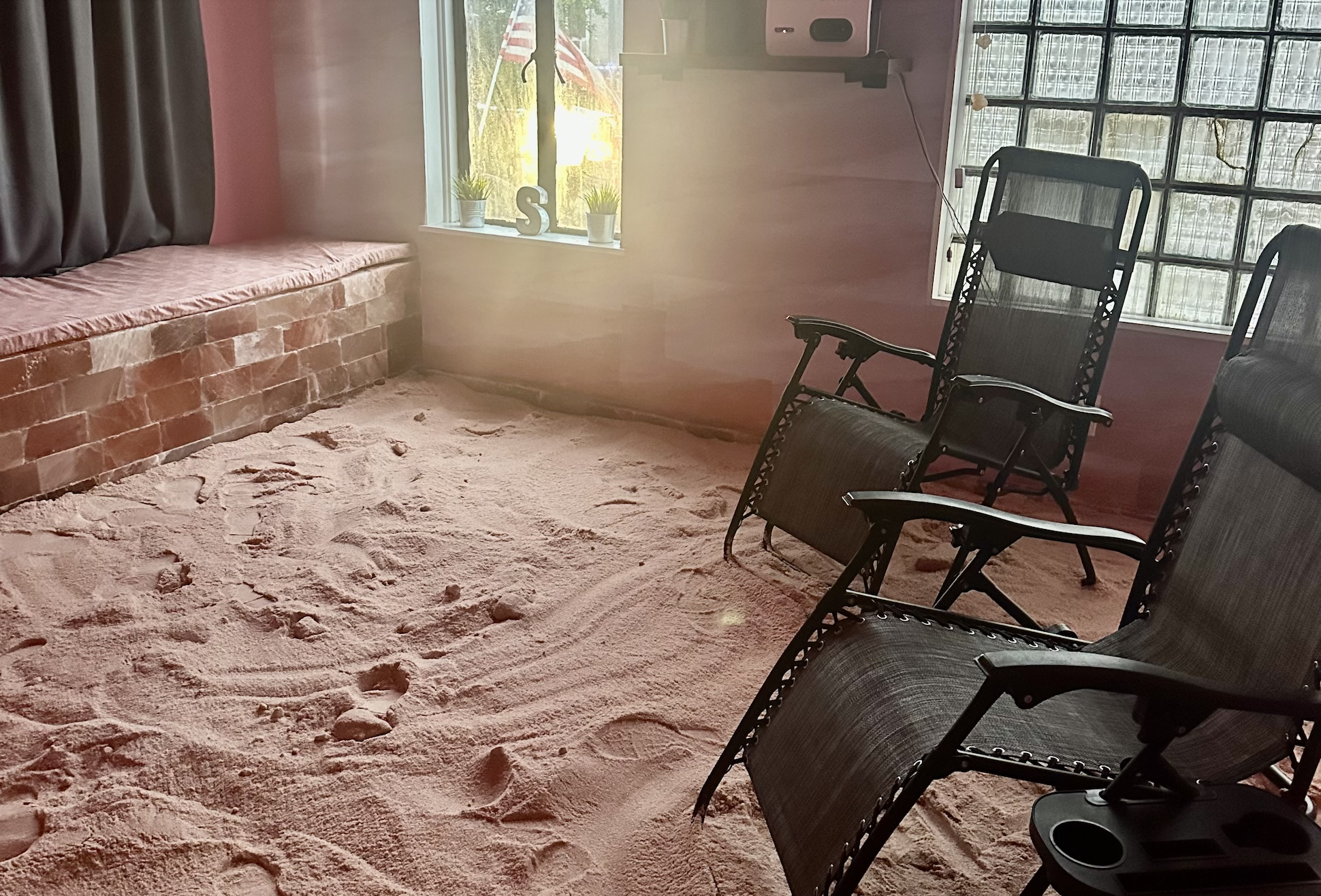 Living room with sand flooring, black chairs, a brick bench under a window with potted plants and decorative items, and sunlight coming through the window.