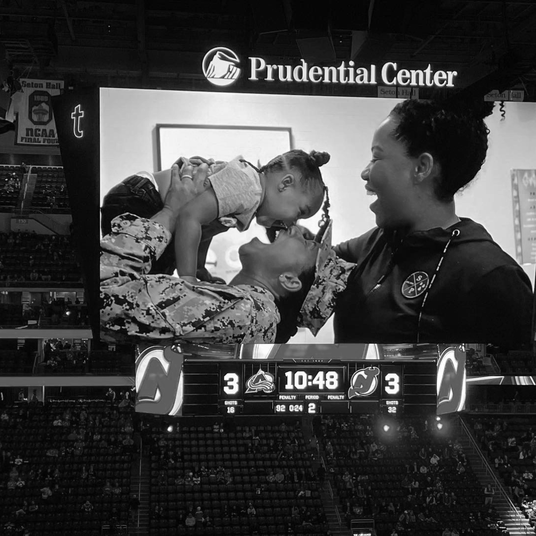 A black and white photograph showing a joyful moment at a sports game. A military service member in uniform is lying down, smiling with a child on their back. The child is leaning over and touching noses with a woman standing nearby, who is also smiling. In the background, a large pizza sign reads "Prudential Center," and a scoreboard shows the game tied at 3-3 with 10 minutes and 48 seconds remaining.