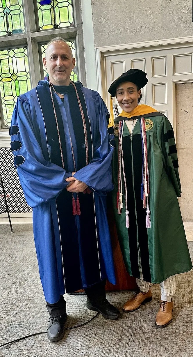 Two people at a graduation ceremony, one in a blue academic gown and the other in a green gown with a hood, both wearing honor cords and tassels, standing indoors near stained glass windows.