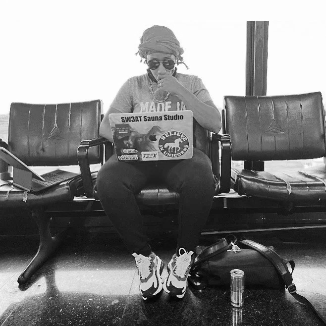 A person sitting on a bench at an airport, using a laptop covered with stickers. They are wearing sunglasses, a headwrap, and casual clothes. There are two bags on the floor near their feet and a water bottle in front of them.