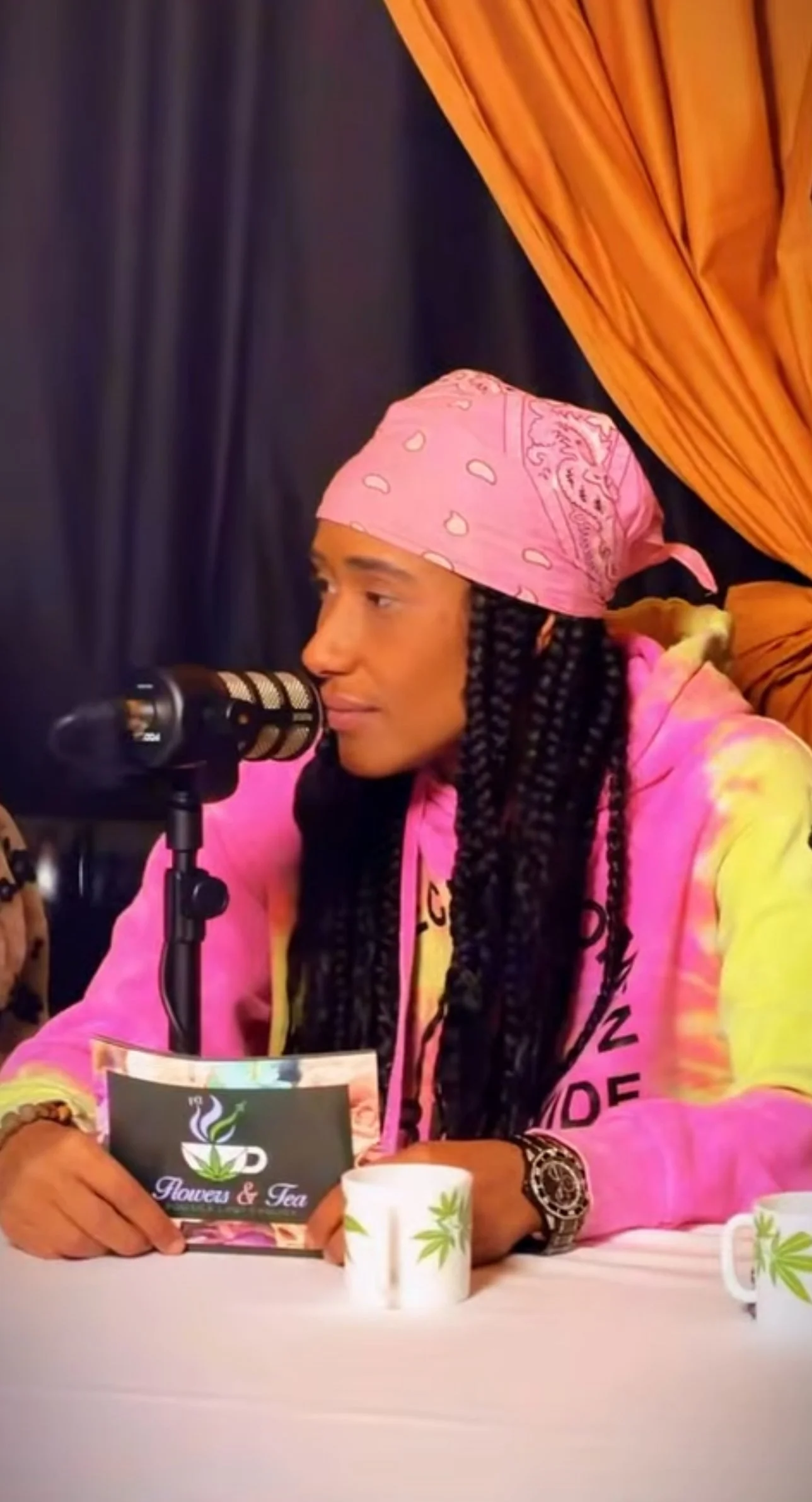 A woman with long braided hair wearing a pink bandana and bright yellow and pink hoodie, sitting at a table with a microphone in front of her. She is holding a flyer with a cannabis leaf logo and the words 'Flowers & Tea,' and there are white coffee mugs with cannabis leaf designs on the table. Behind her are dark blue and orange curtains.