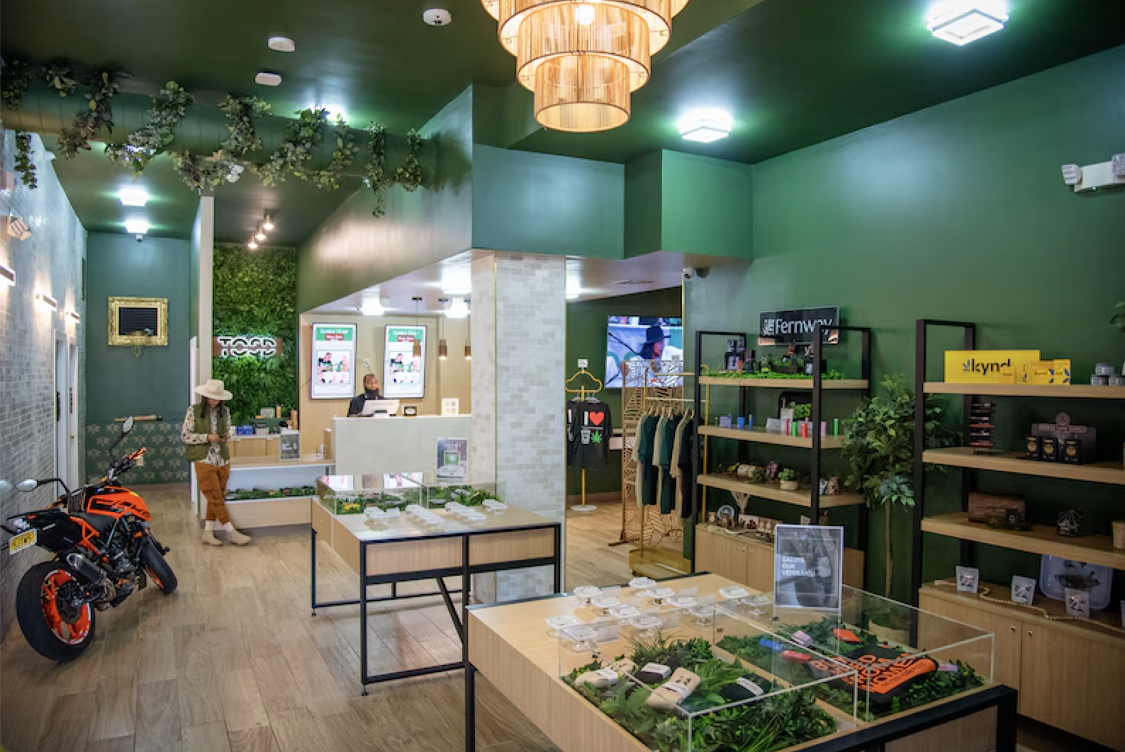 Interior view of a modern retail store with green walls, wooden shelves, and display tables. A motorcycle is parked inside, and a customer is browsing near the checkout counter. The store has decorative lighting, clothing racks, and various products 