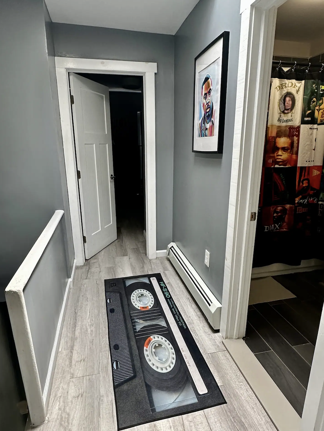 Hallway with a cassette tape rug, open door to a dark room, wall art, and a doorway to a room with curtain featuring rapper images.