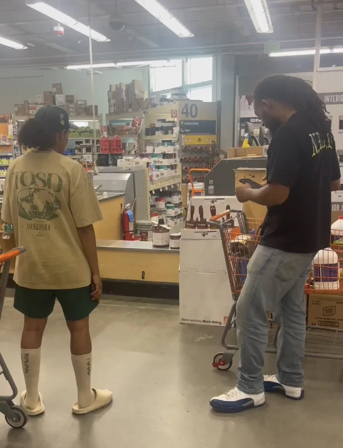 Two young men standing at a checkout counter in a store, one is wearing a beige TOSD t-shirt, the other a black TOSD t-shirt, both with long shorts and sneakers. The store has shelves stocked with various products, and a shopping cart is visible.