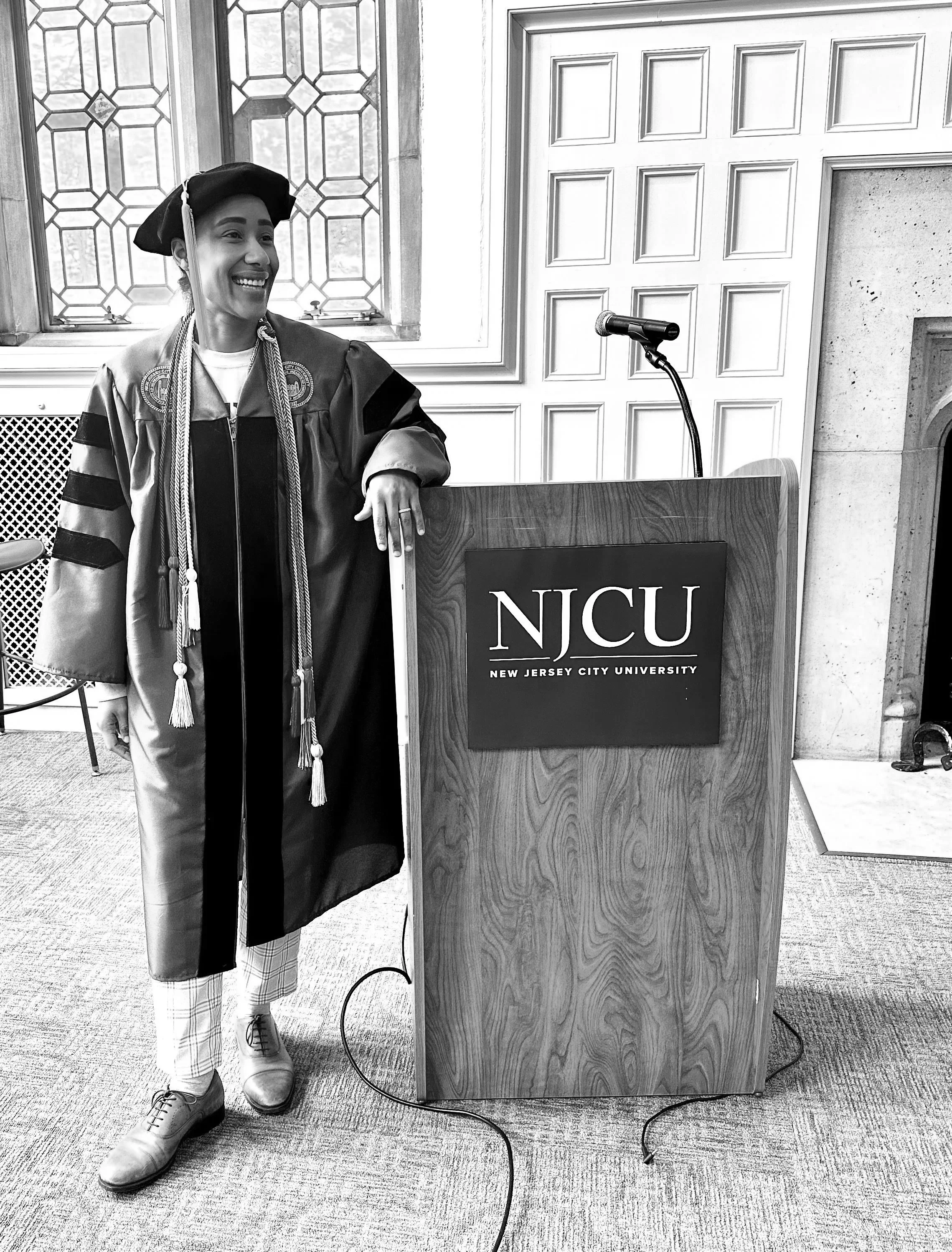Woman in graduation gown and cap standing next to podium with NJCU logo, smiling in a decorated room. Dr Beast at New Jersey City University in Jersey City