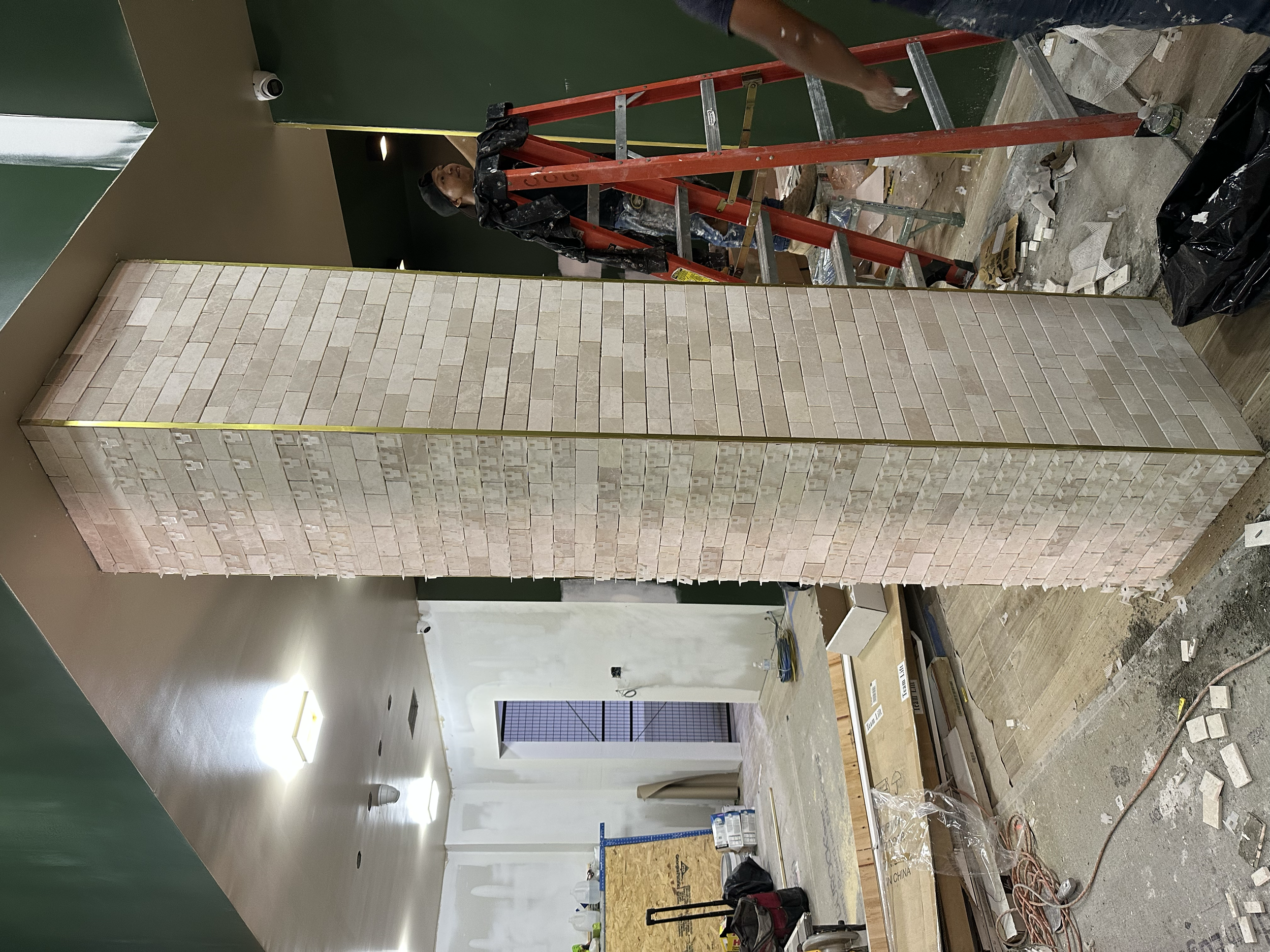 Construction workers installing a beige brick wall on a construction site. One worker is standing on a red ladder, working on the top of the wall. The area is cluttered with construction materials, tools, and debris.