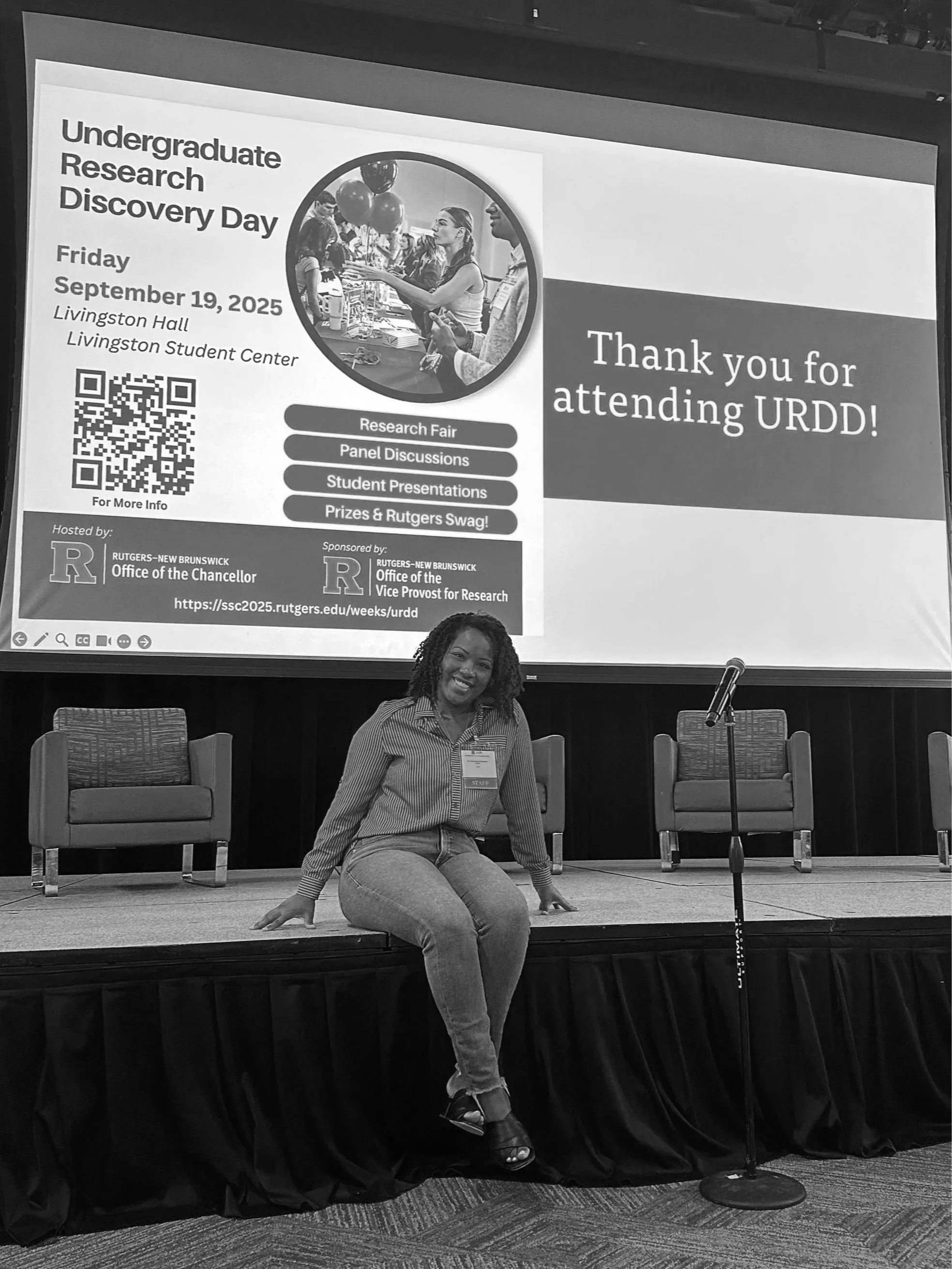 A woman sitting on the edge of a stage in a conference room, smiling at the camera, with a large screen behind her displaying information about Undergraduate Research Discovery Day at Rutgers University, including a QR code, event details, and a message thanking attendees.