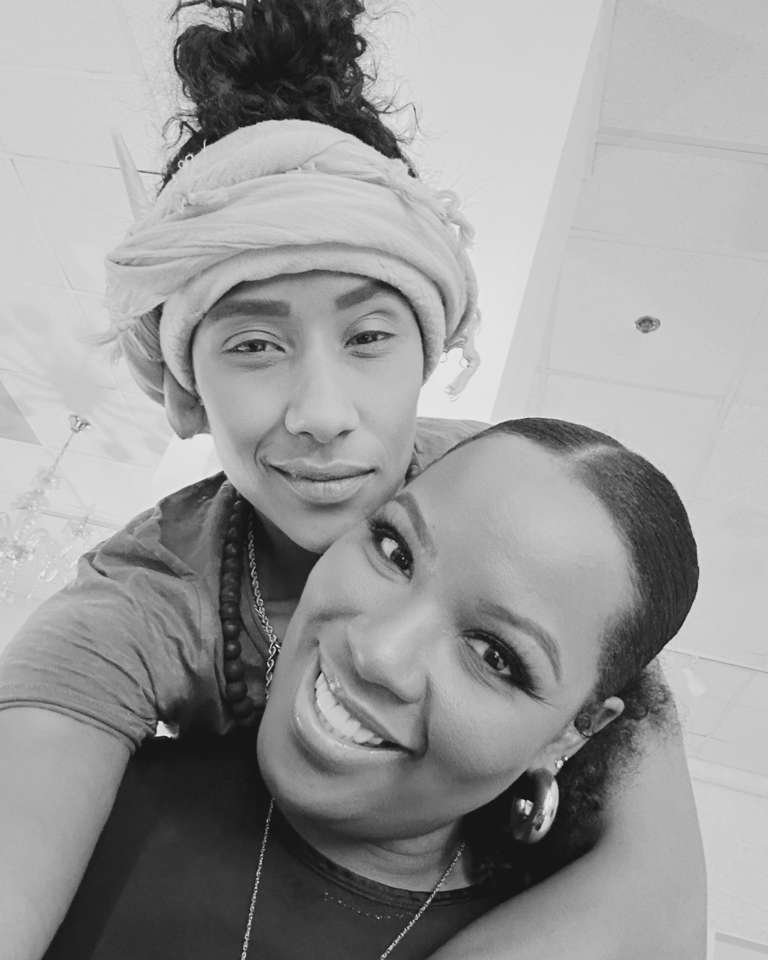 Two women smiling (Dr(s). Brevard-Rodriguez, and posing for a selfie in an indoor setting, with one woman wearing headwraps and the other having short curly hair.