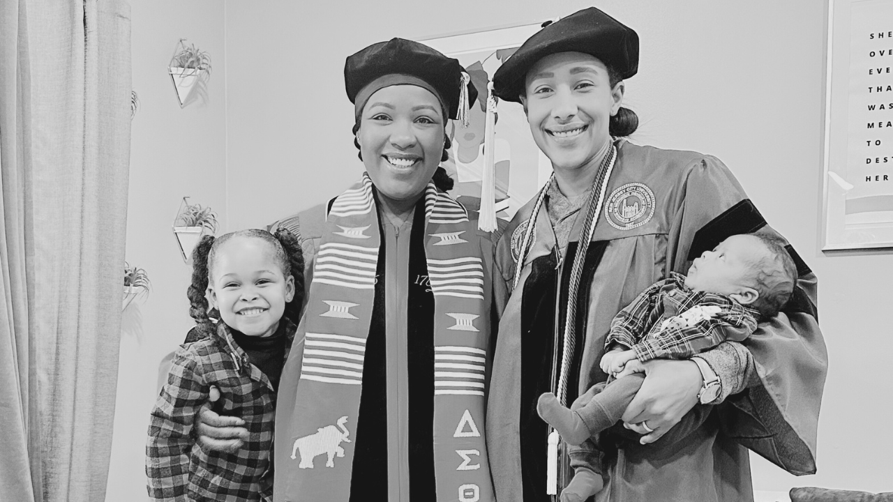 A woman and a man in graduation caps and gowns are smiling and posing with two children. The woman is wearing a sash and the man is holding a baby, while a young girl hugs the woman.