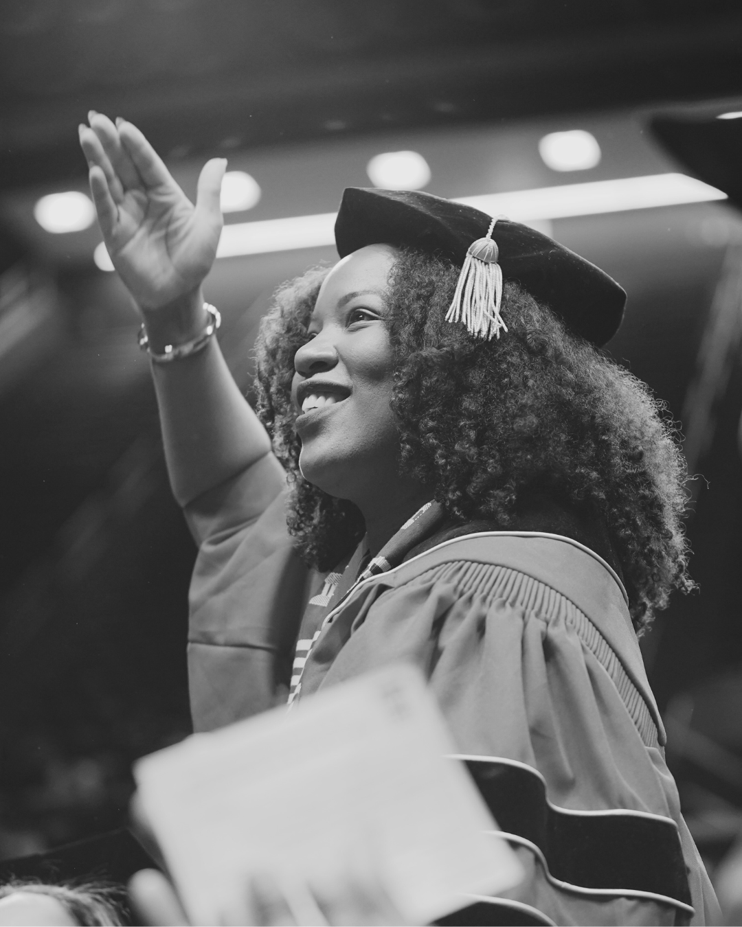 A young woman with curly hair wearing a graduation cap and gown, smiling and waving. At Rutgers University