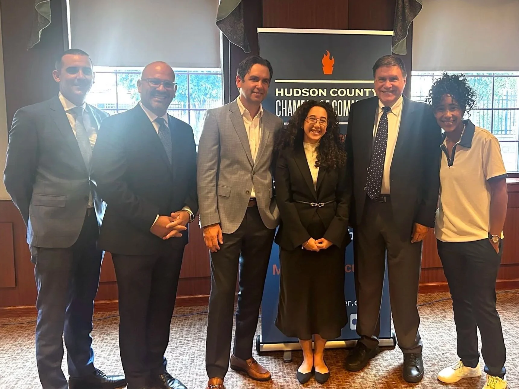 Group of six professionals standing together indoors in front of a Hudson County Chamber of Commerce banner.