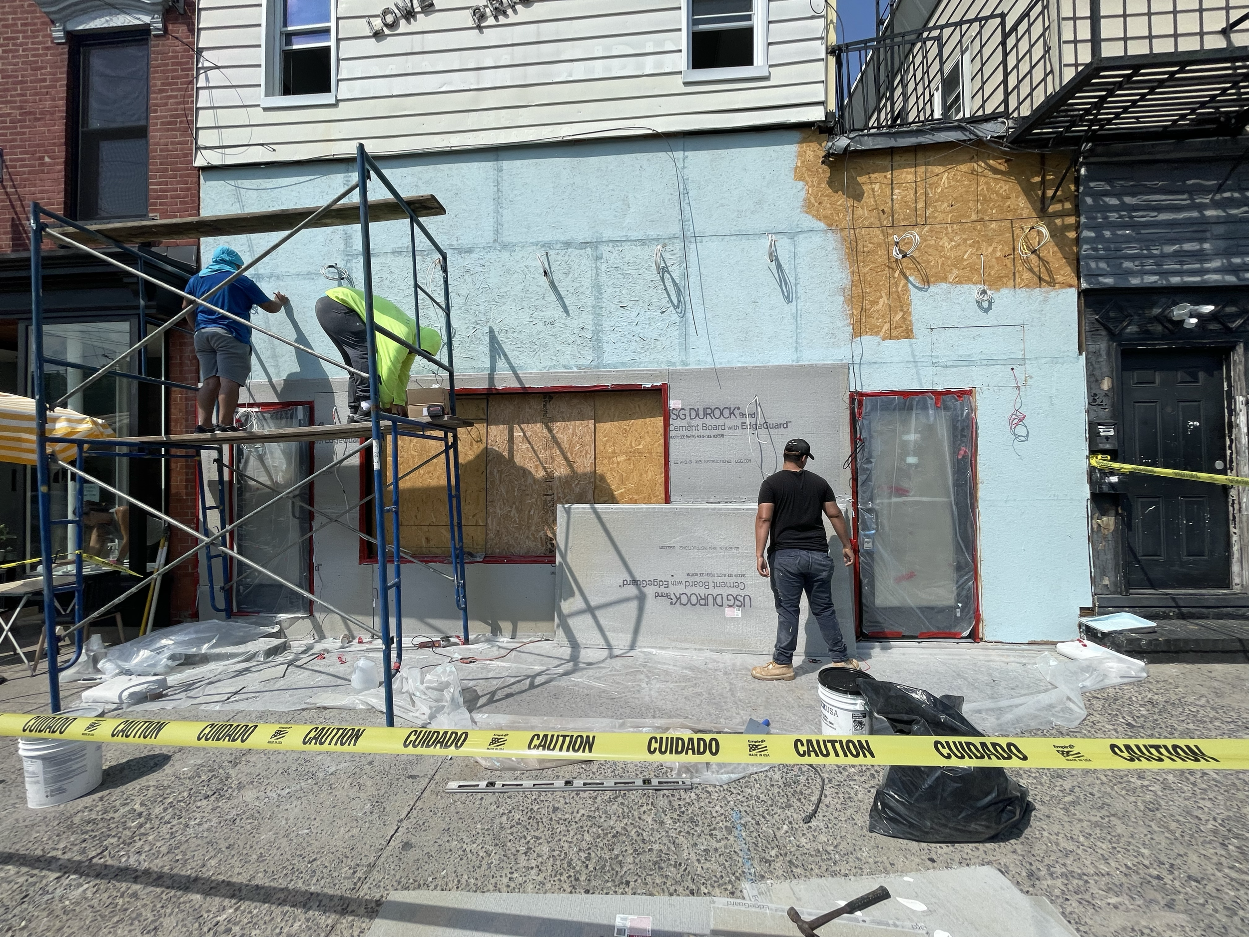 Construction workers painting the exterior wall of a building, with caution tape in the foreground and construction materials on the ground.
